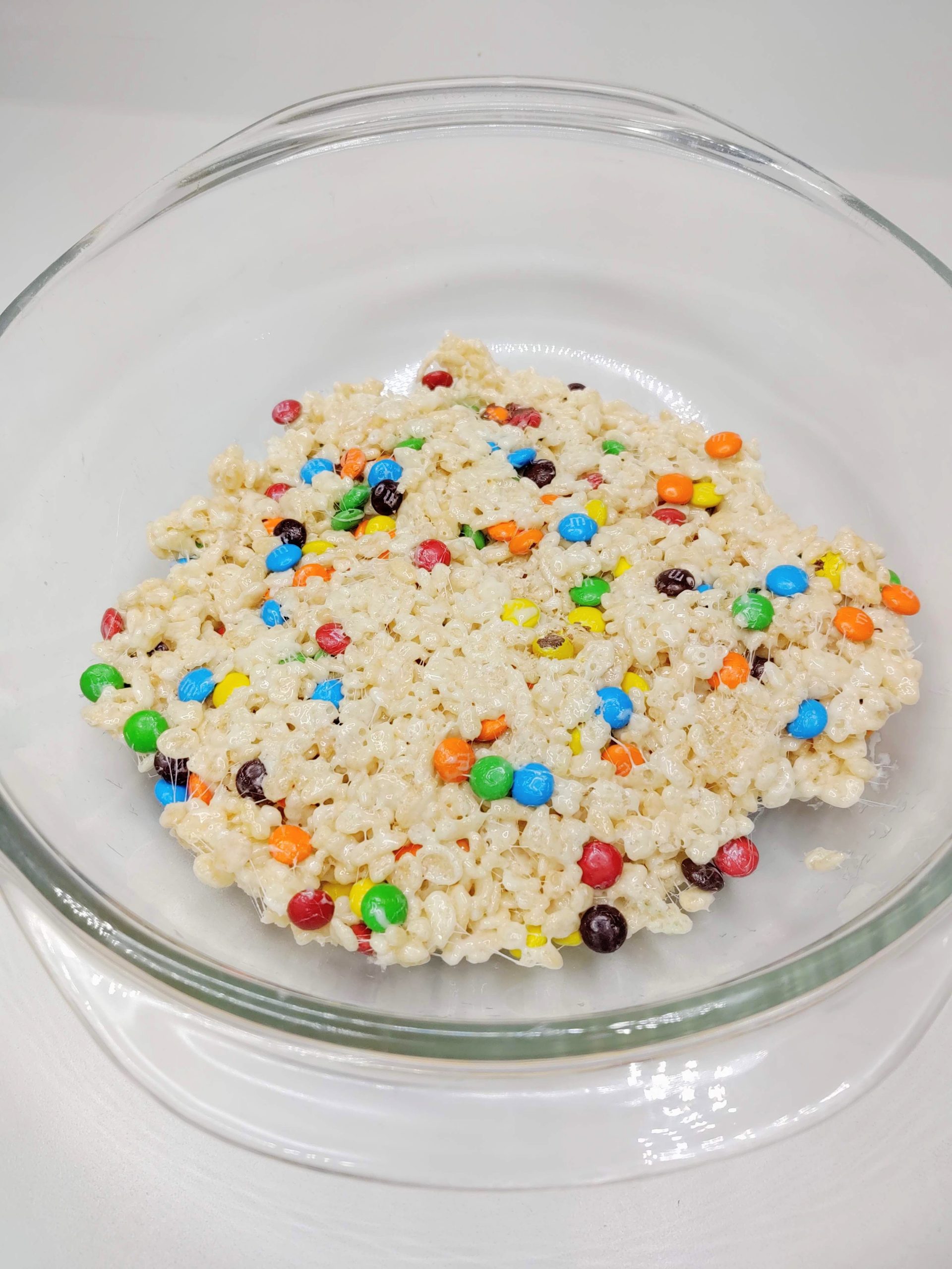 M&Ms being mixed with marshmallows and cereal in a glass bowl