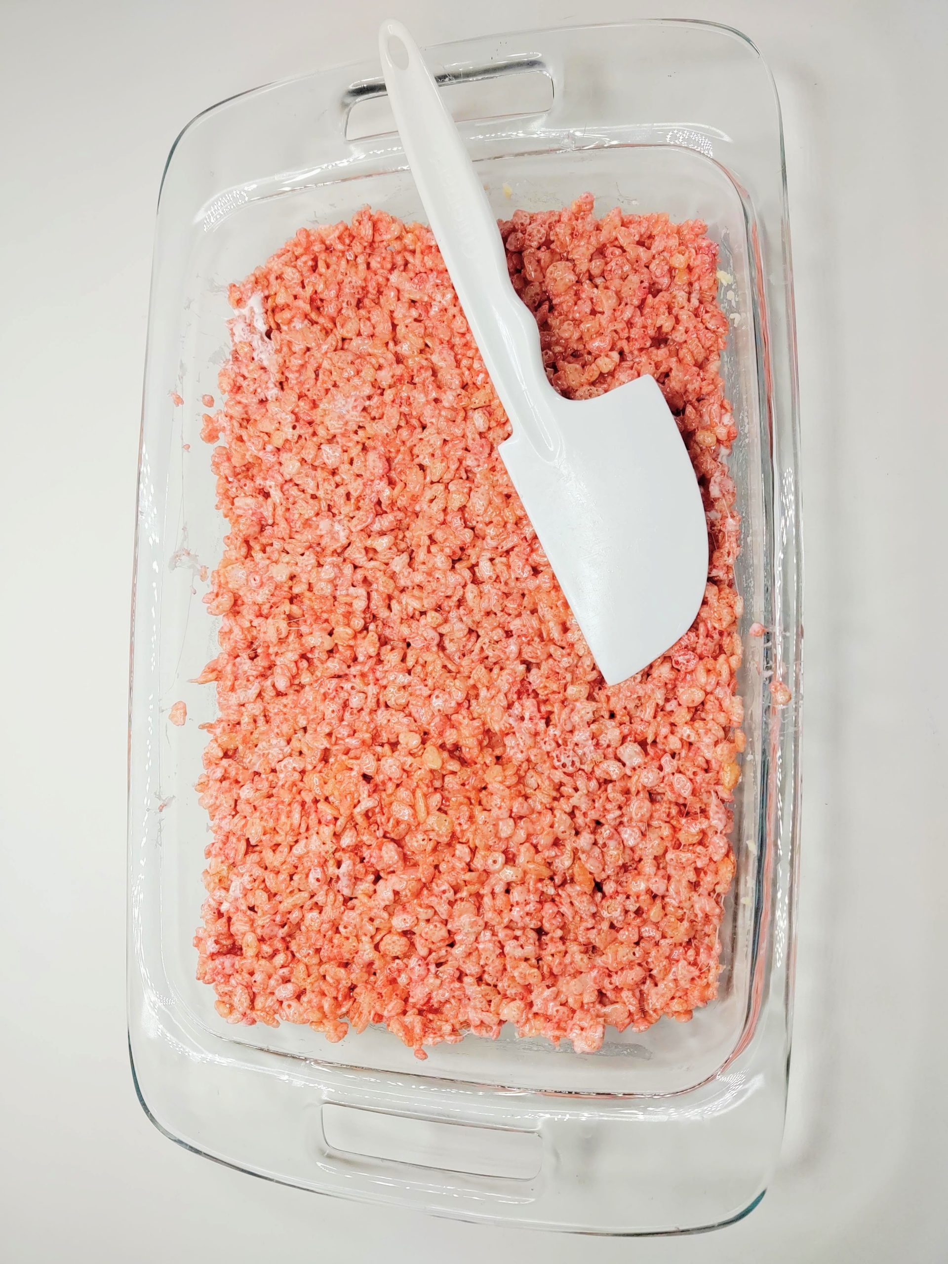 marshmallow and cereal mixture pressed into glass pan with spatula on top