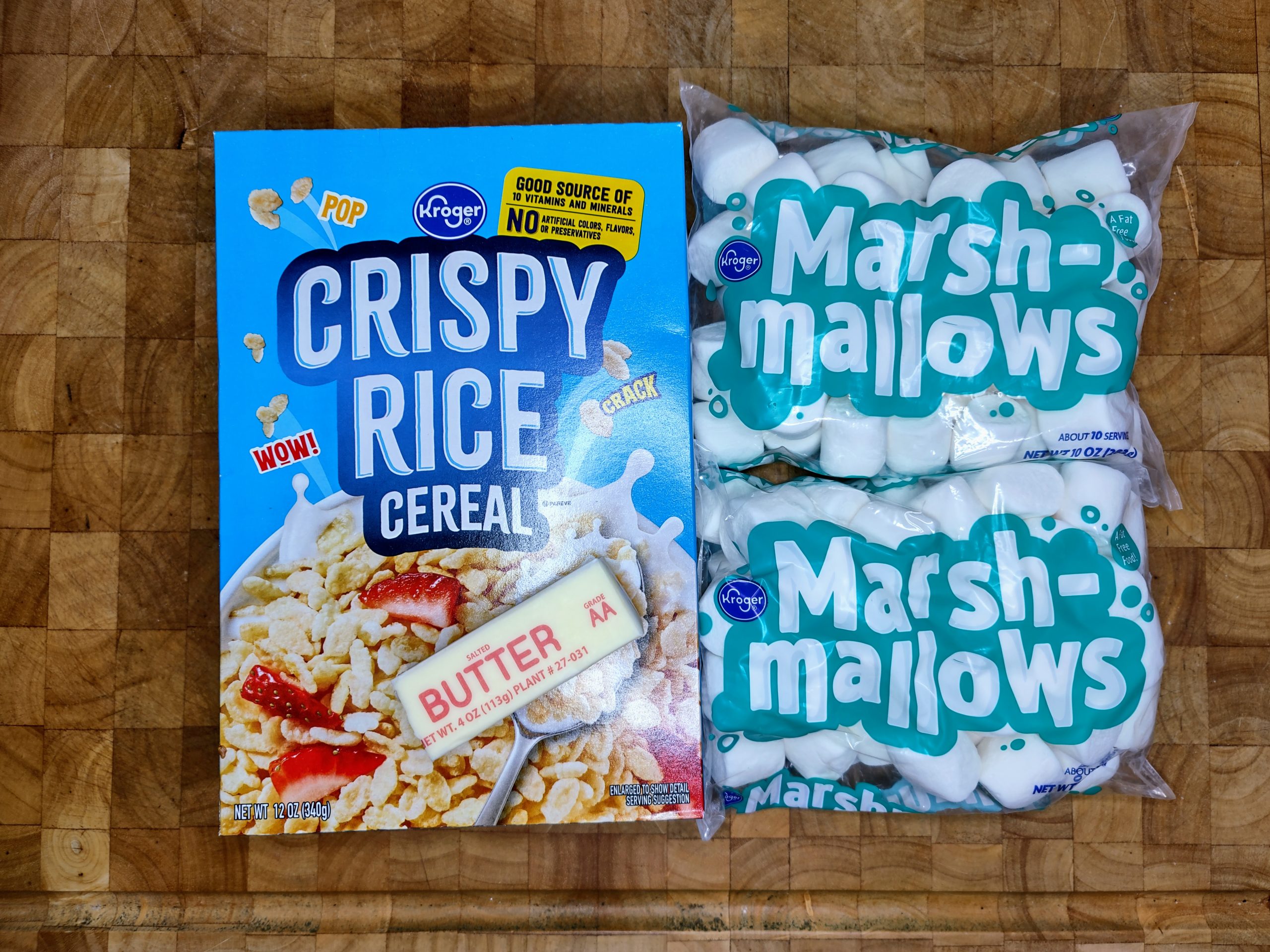 two bags of marshmallows, stick of butter and box of rice crispy cereal on a wooden table