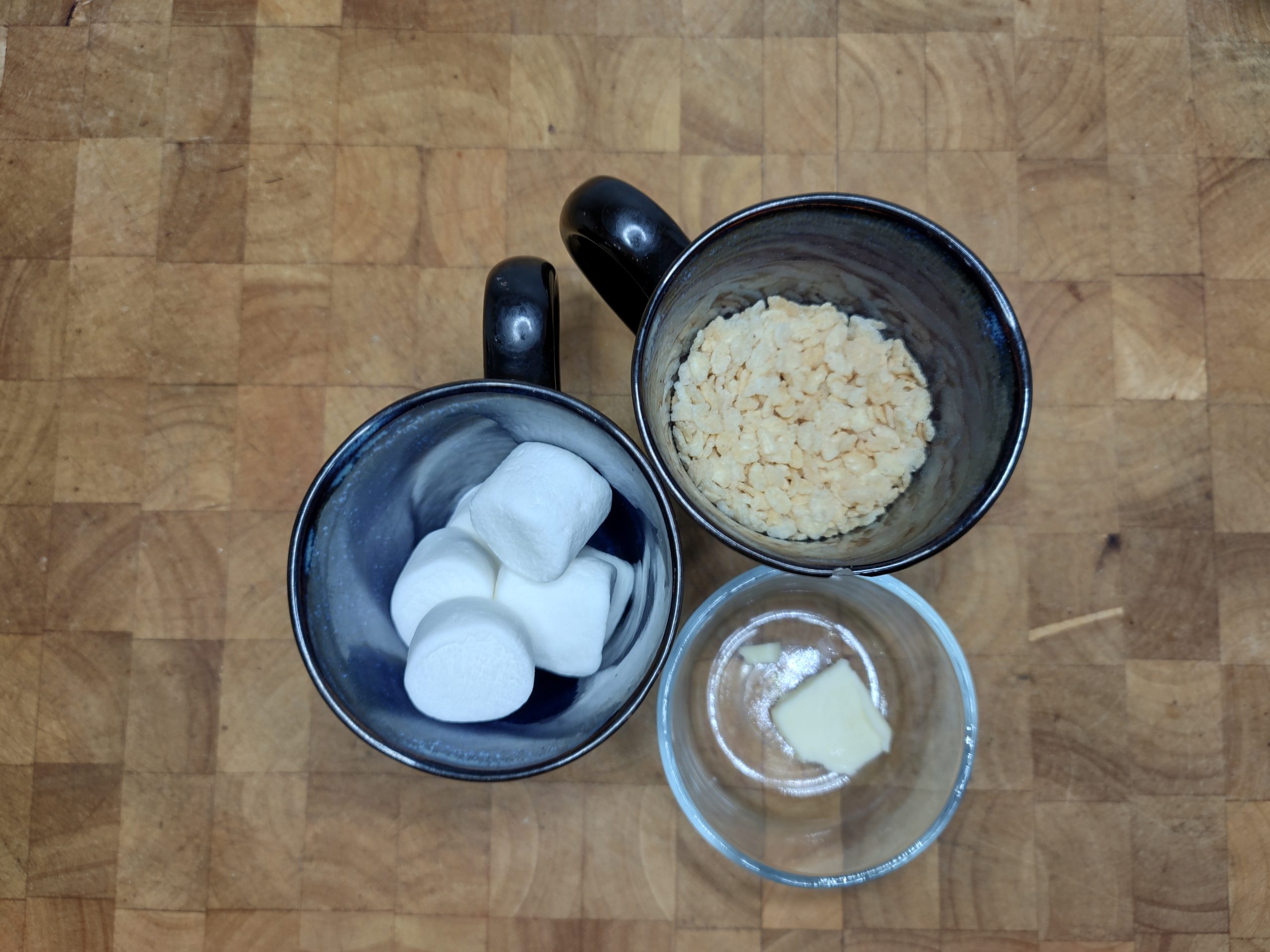 mug of marshmallows, mug of rice krispie cereal and bowl of butter on wooden table