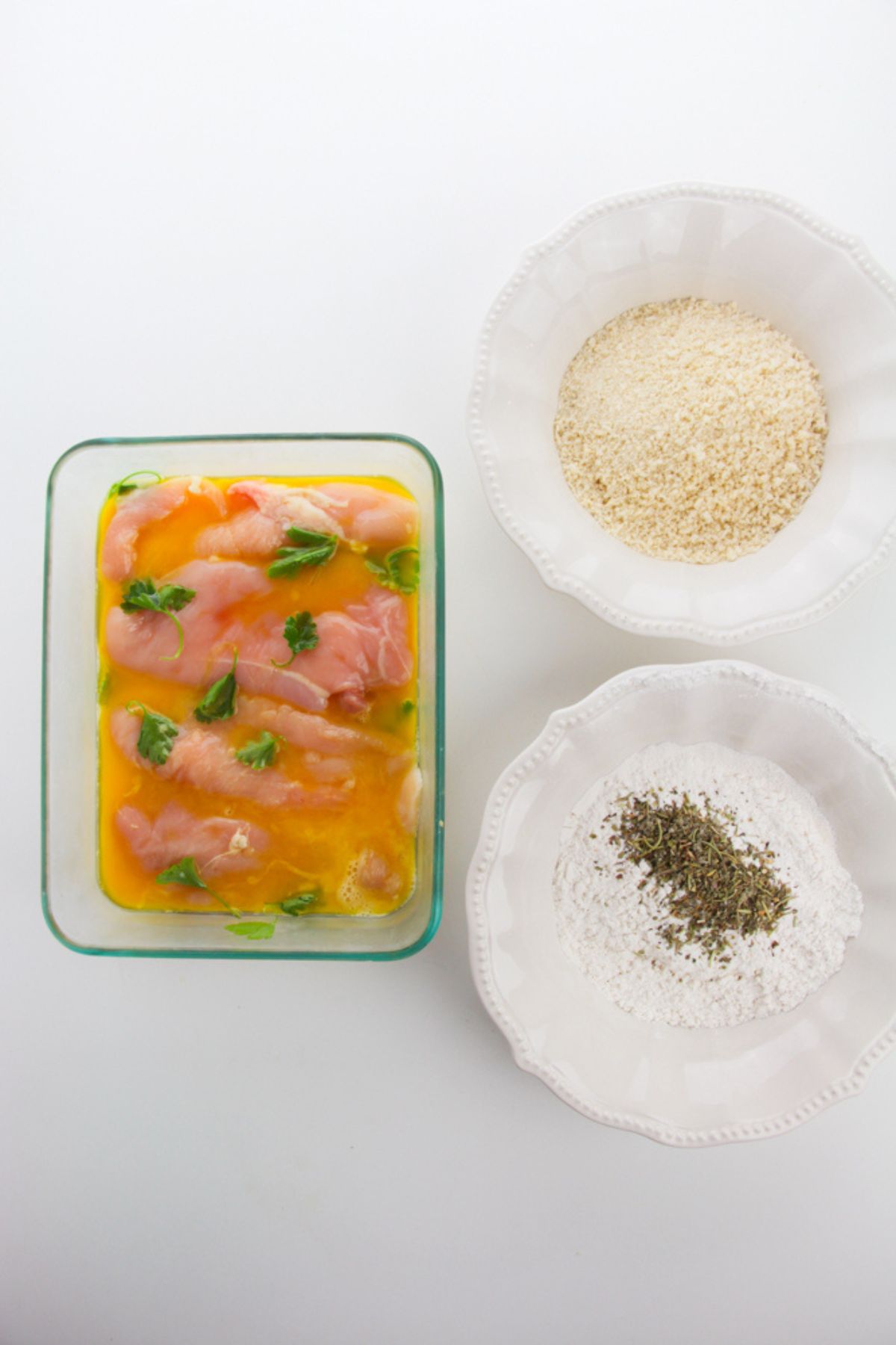 Chicken in egg mixture next to bowls of flour and spices.