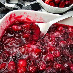 Homemade cranberry sauce in a white baking dish with a spoon, fresh cranberries in a small bowl on a black and white plaid...