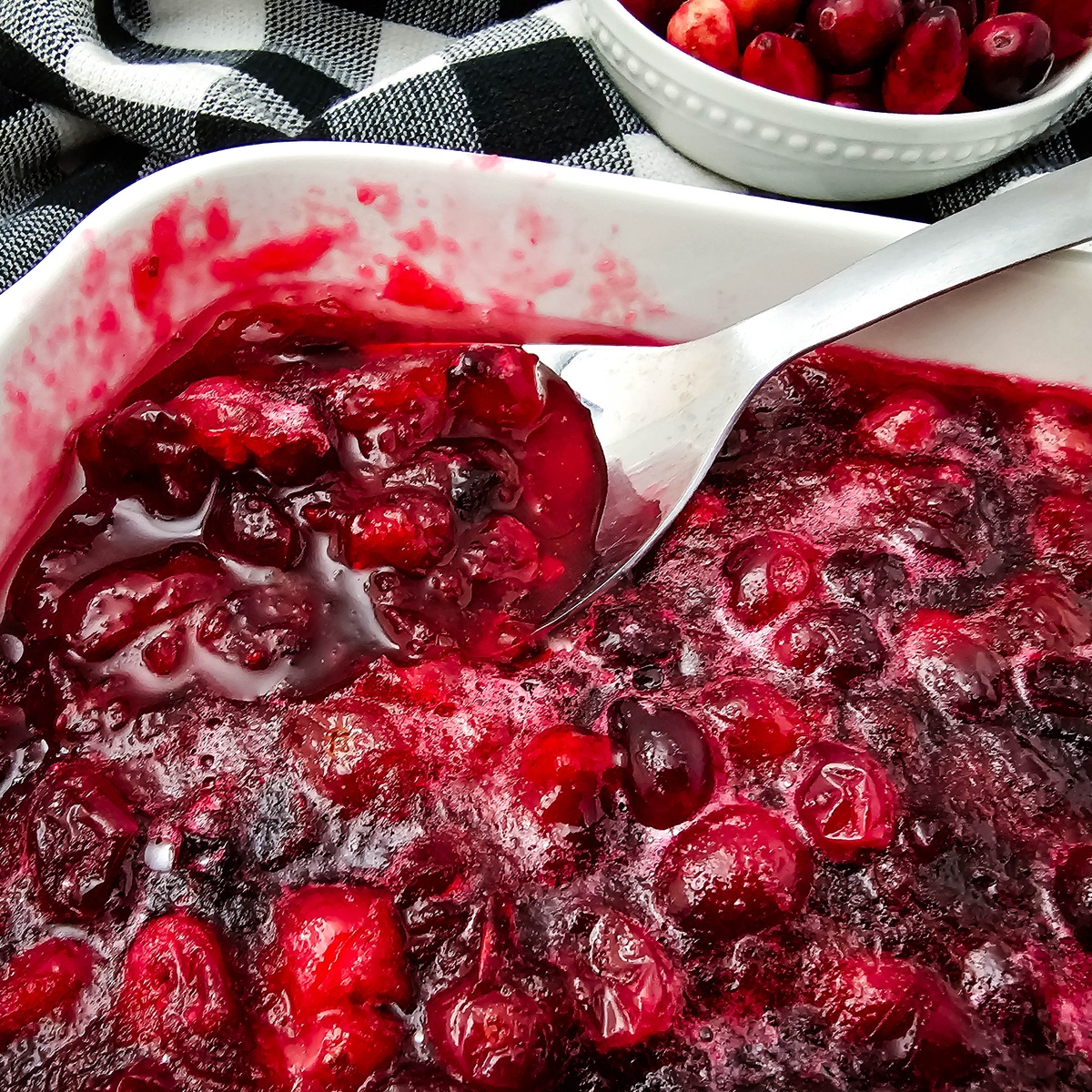 Homemade cranberry sauce in a white baking dish with a spoon, fresh cranberries in a small bowl on a black and white plaid...