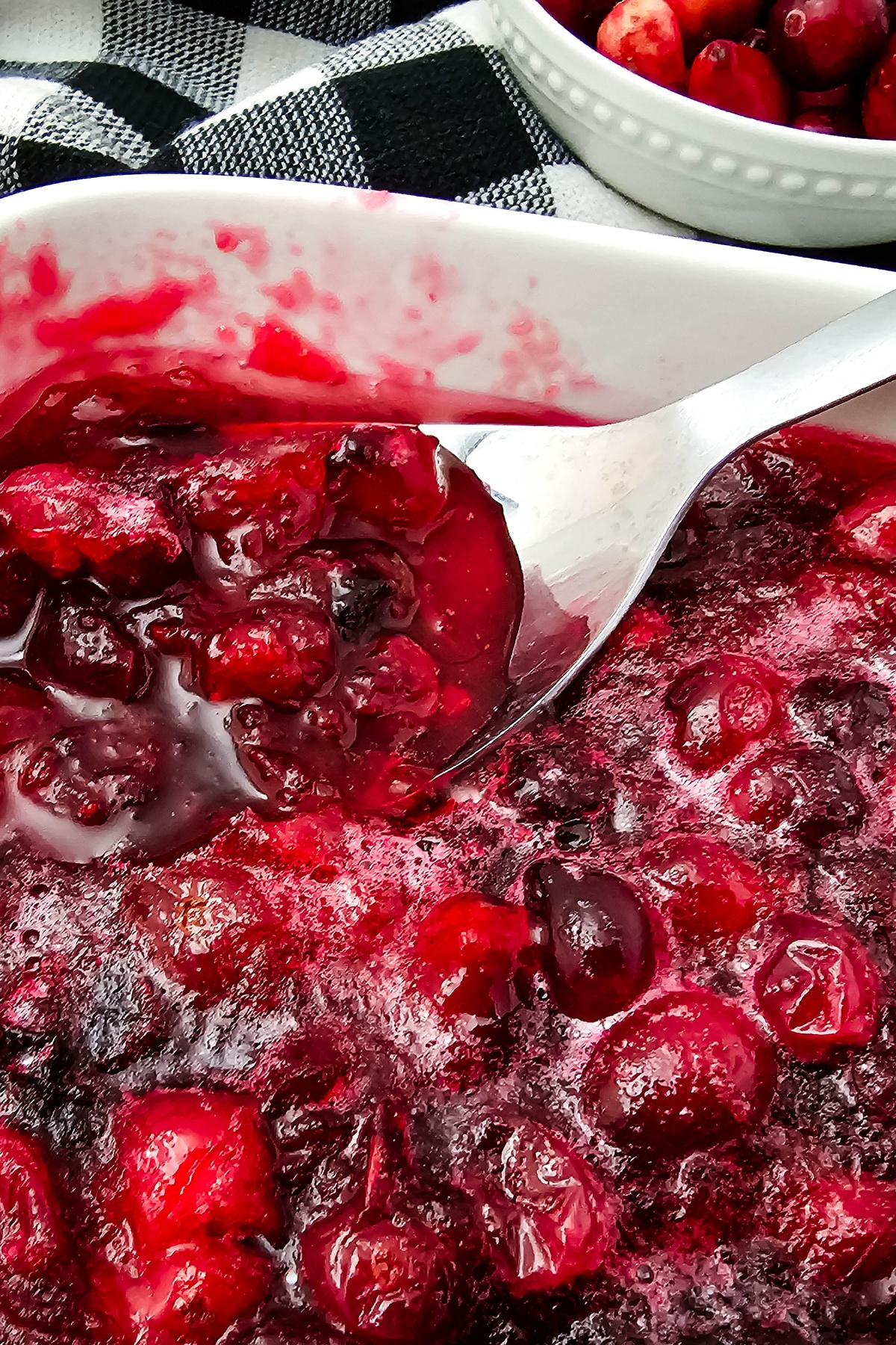 A spoon scoops glossy cranberry sauce from a white serving dish, with fresh cranberries in a bowl on a black and white pla...