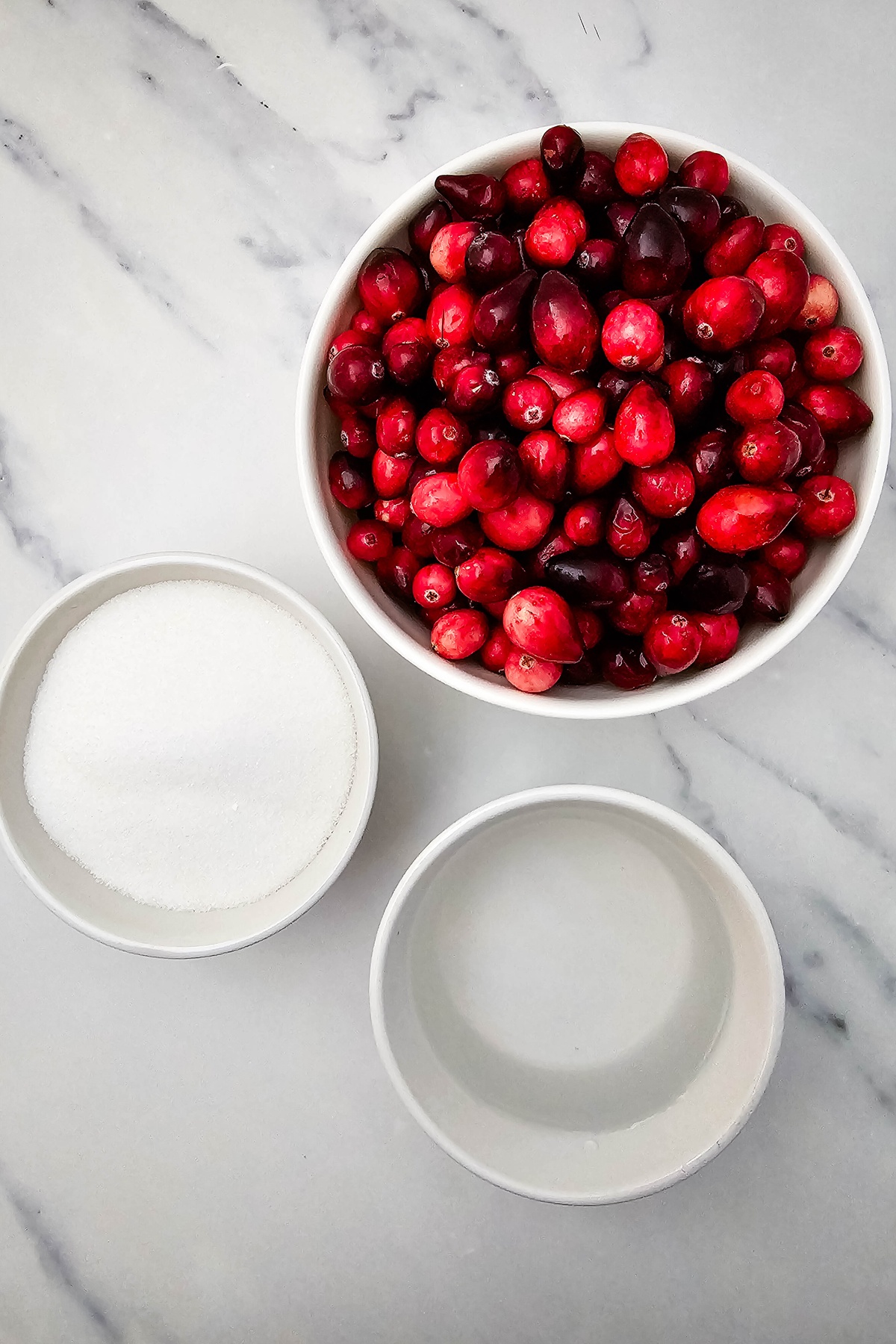 Three white bowls on marble surface containing fresh raw cranberries, white sugar, and water.