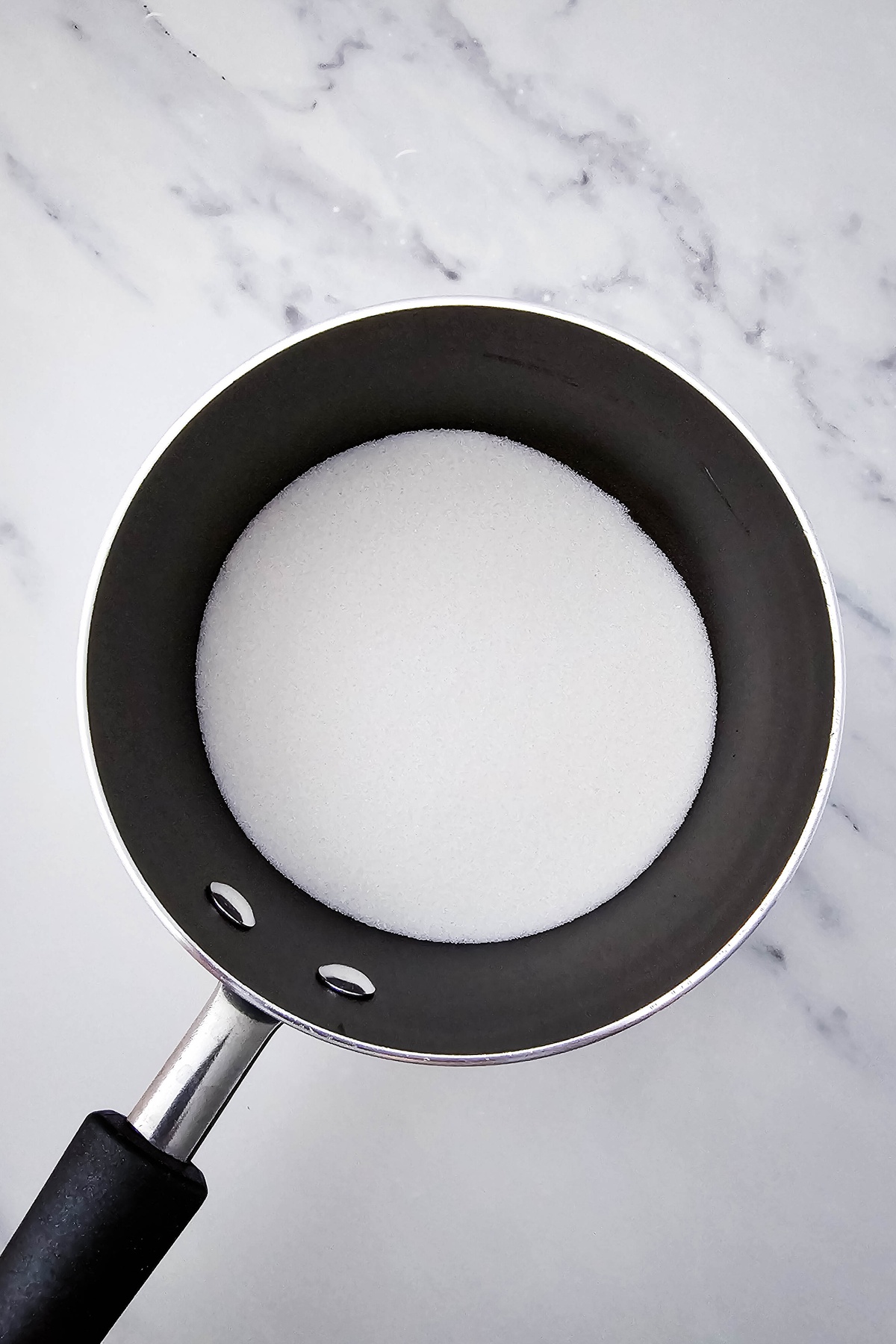 White granulated sugar in a dark nonstick saucepan on a marble surface, ready to be heated.