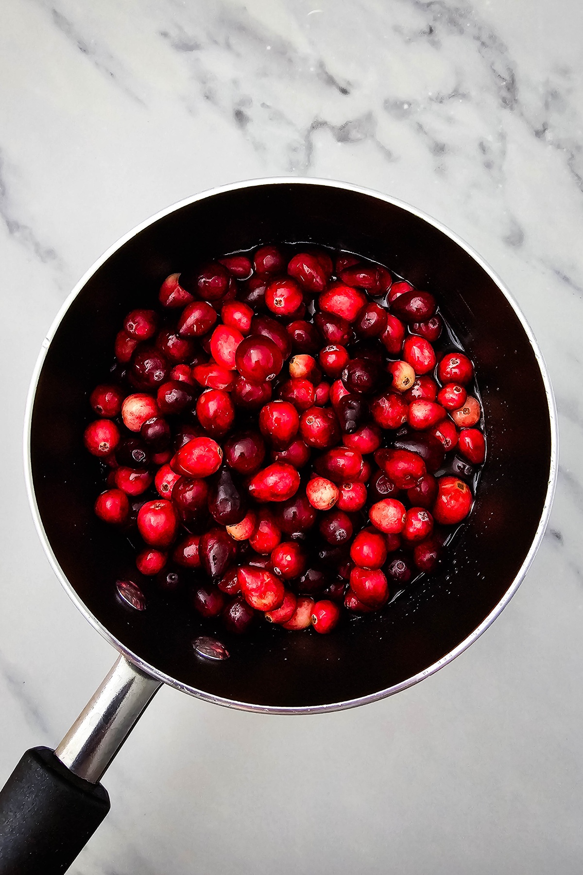 Fresh cranberries in dark liquid in a saucepan on a marble surface, ready to cook