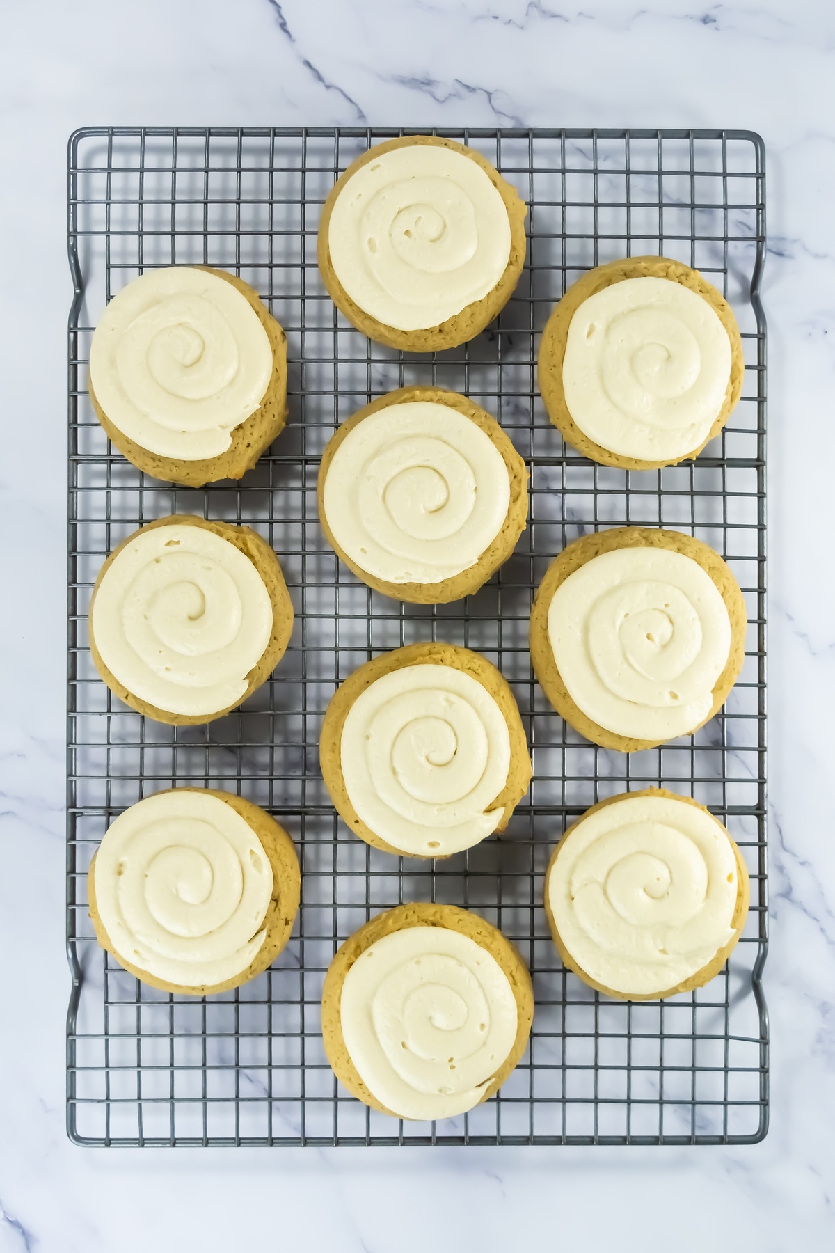Ten frosted pumpkin cookies with white cream cheese frosting piped in spiral patterns cooling on a wire rack.