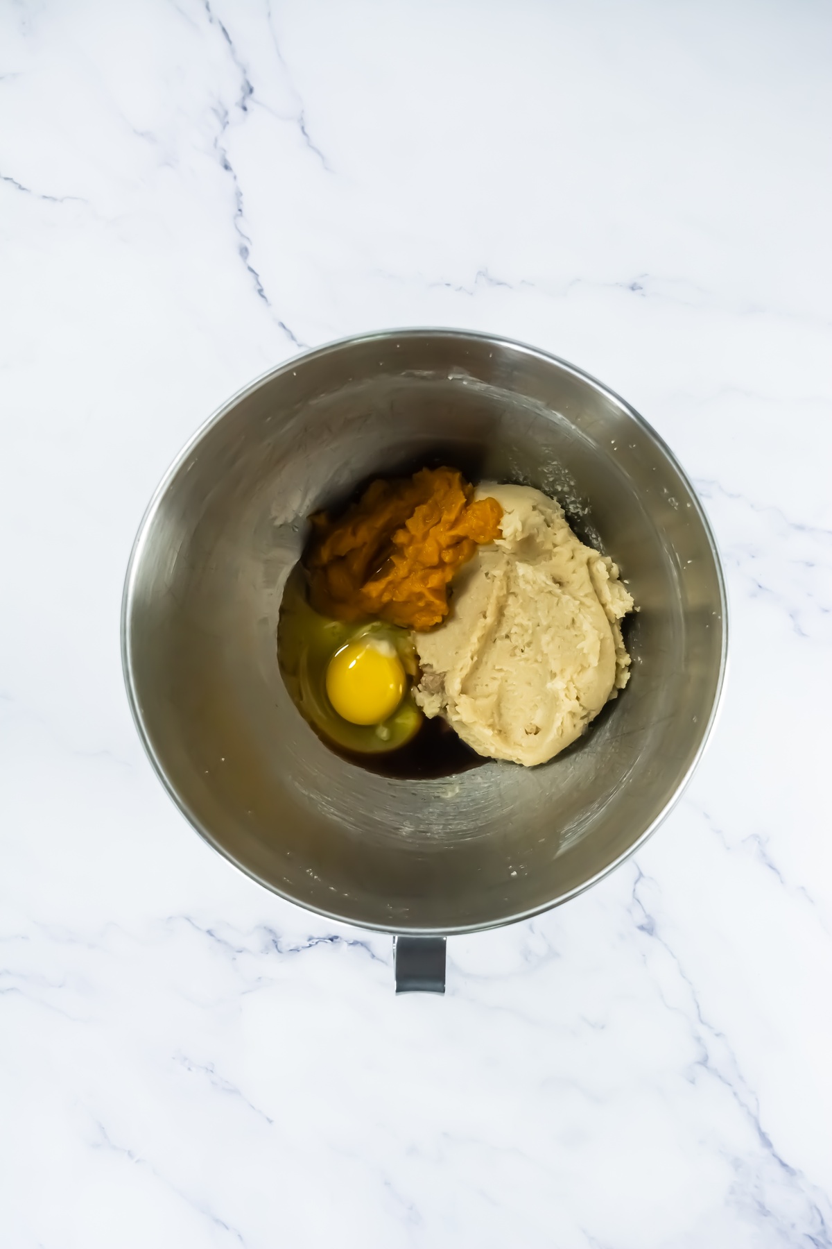 Stainless steel mixing bowl containing creamed butter and sugar, pumpkin puree, a raw egg, and vanilla extract before mixing