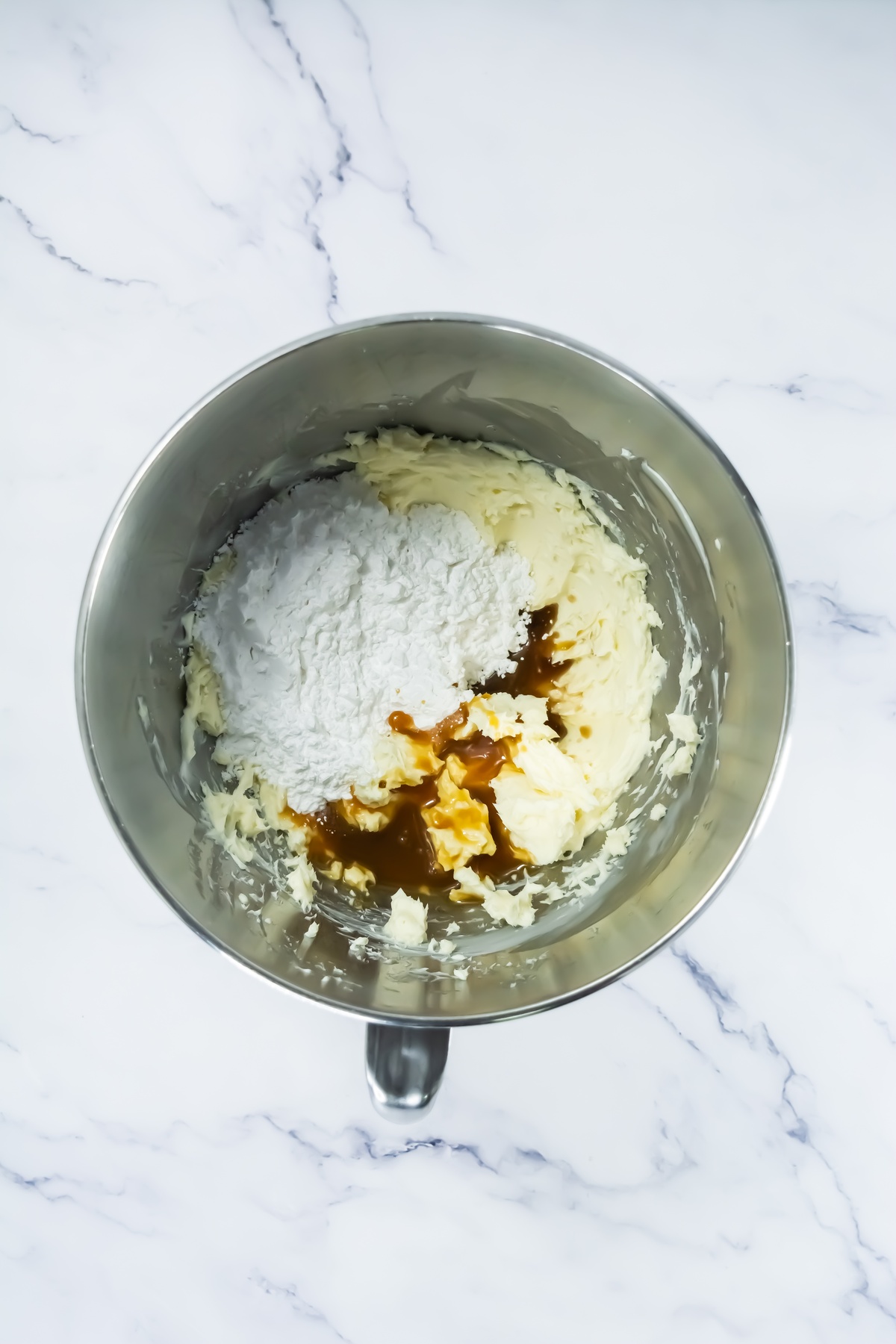 Stainless steel mixing bowl containing whipped cream cheese, powdered sugar, and caramel sauce before mixing.