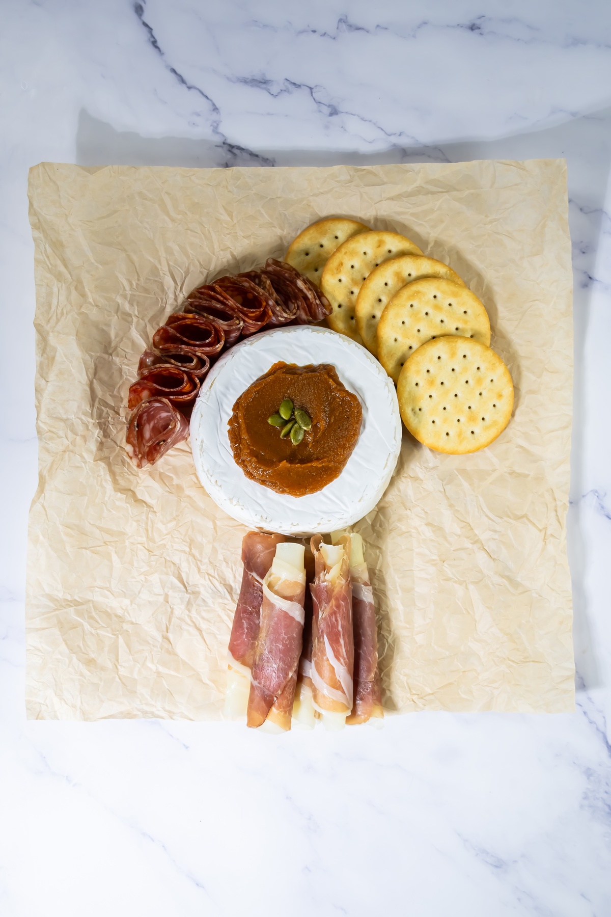 Brie wheel topped with pumpkin butter and pepitas, surrounded by rolled salami, crackers, and prosciutto-wrapped cheese.