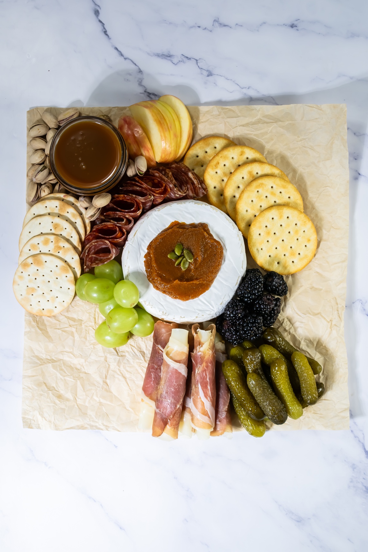 Fall charcuterie board with brie topped with pumpkin butter, crackers, prosciutto-wrapped cheese, salami, grapes, blackberries.