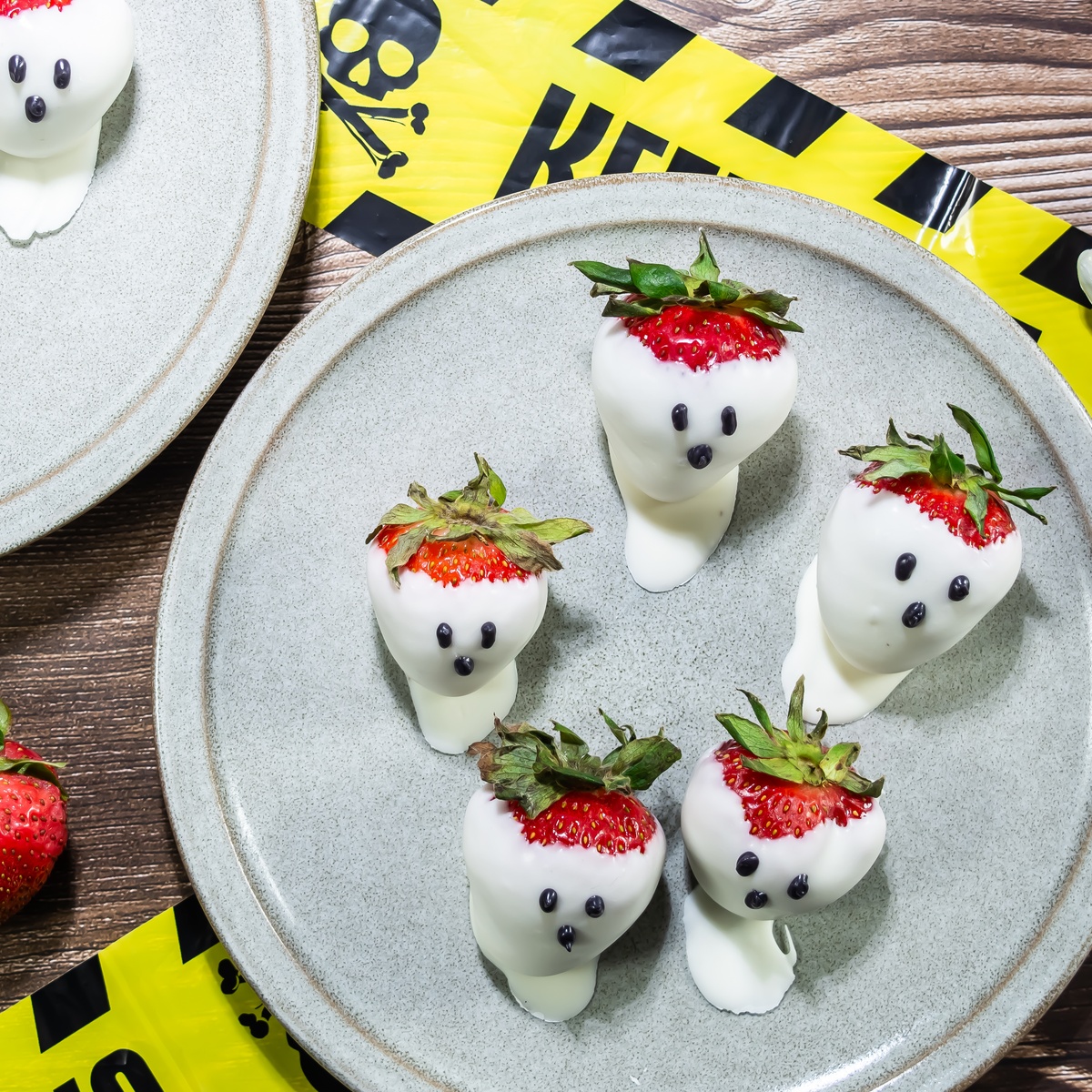 White chocolate-dipped strawberries decorated as ghosts with chocolate dot faces on gray plates with yellow caution tape.