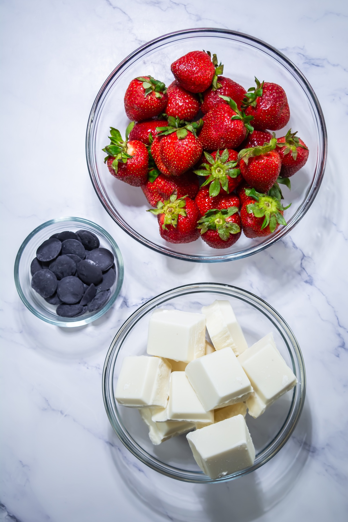Fresh whole strawberries, white chocolate cubes, and dark chocolate wafers in glass bowls on marble surface.