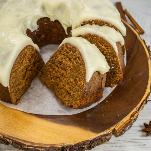 Sliced gingerbread bundt cake topped with cream cheese frosting on a rustic wooden board with cinnamon sticks and star anise.