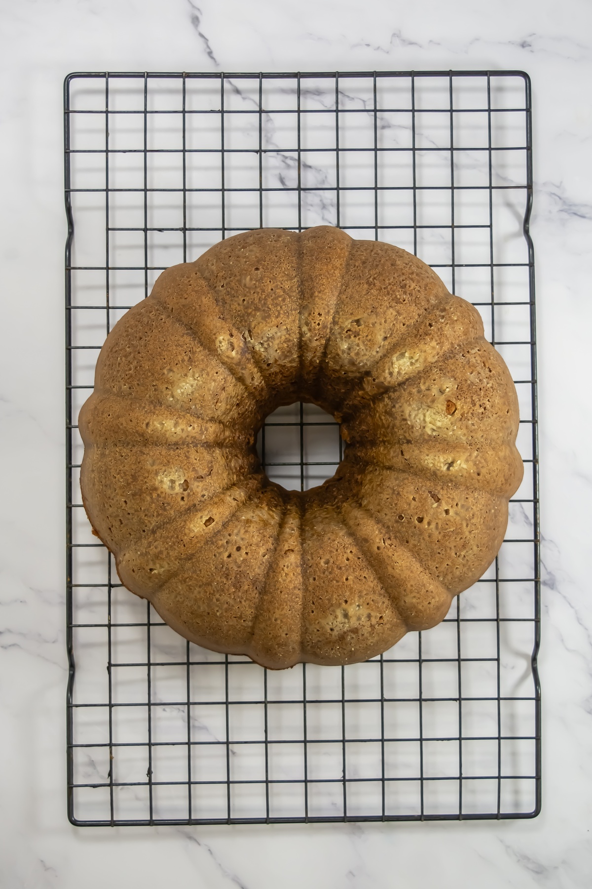 Golden-brown baked bundt cake cooling on a black wire rack on a white marble surface.