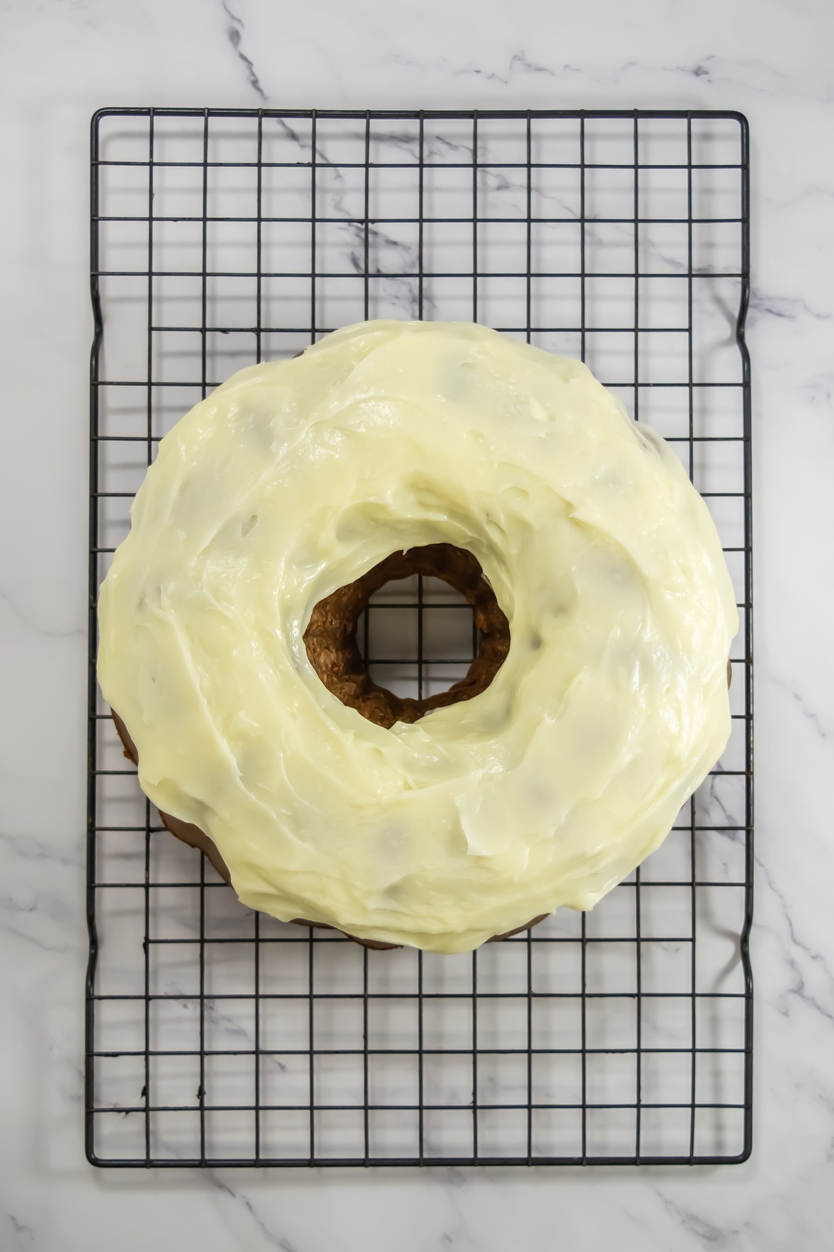 Baked bundt cake topped with cream cheese frosting on a black wire cooling rack on marble surface.