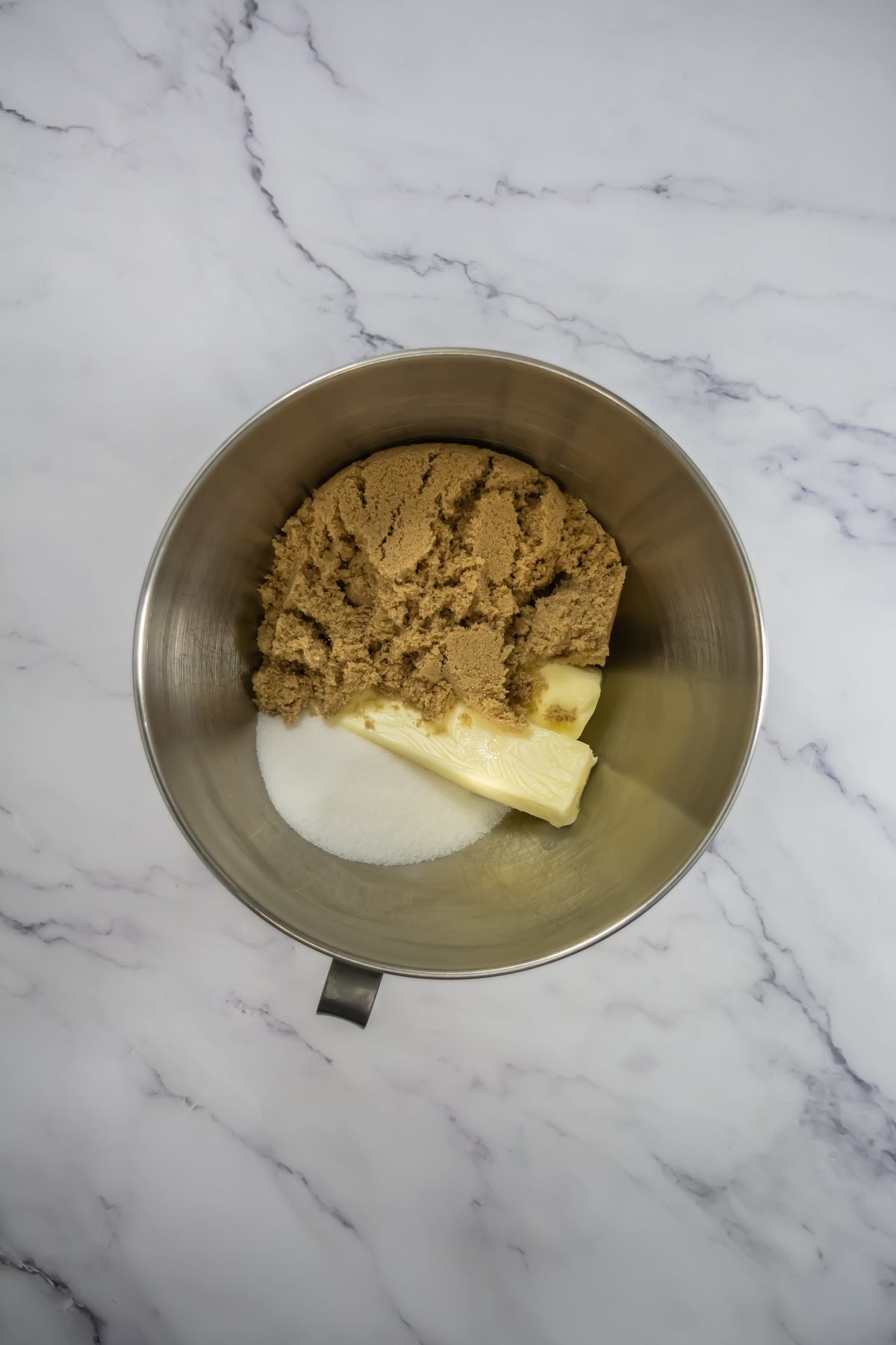 Brown sugar, white sugar, and butter in a stainless steel mixing bowl on a marble surface, ready to be creamed together.