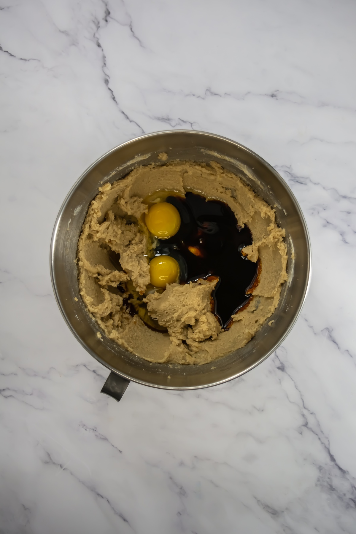 Stainless steel mixing bowl with creamed butter mixture, two raw eggs, and vanilla on a white marble surface.