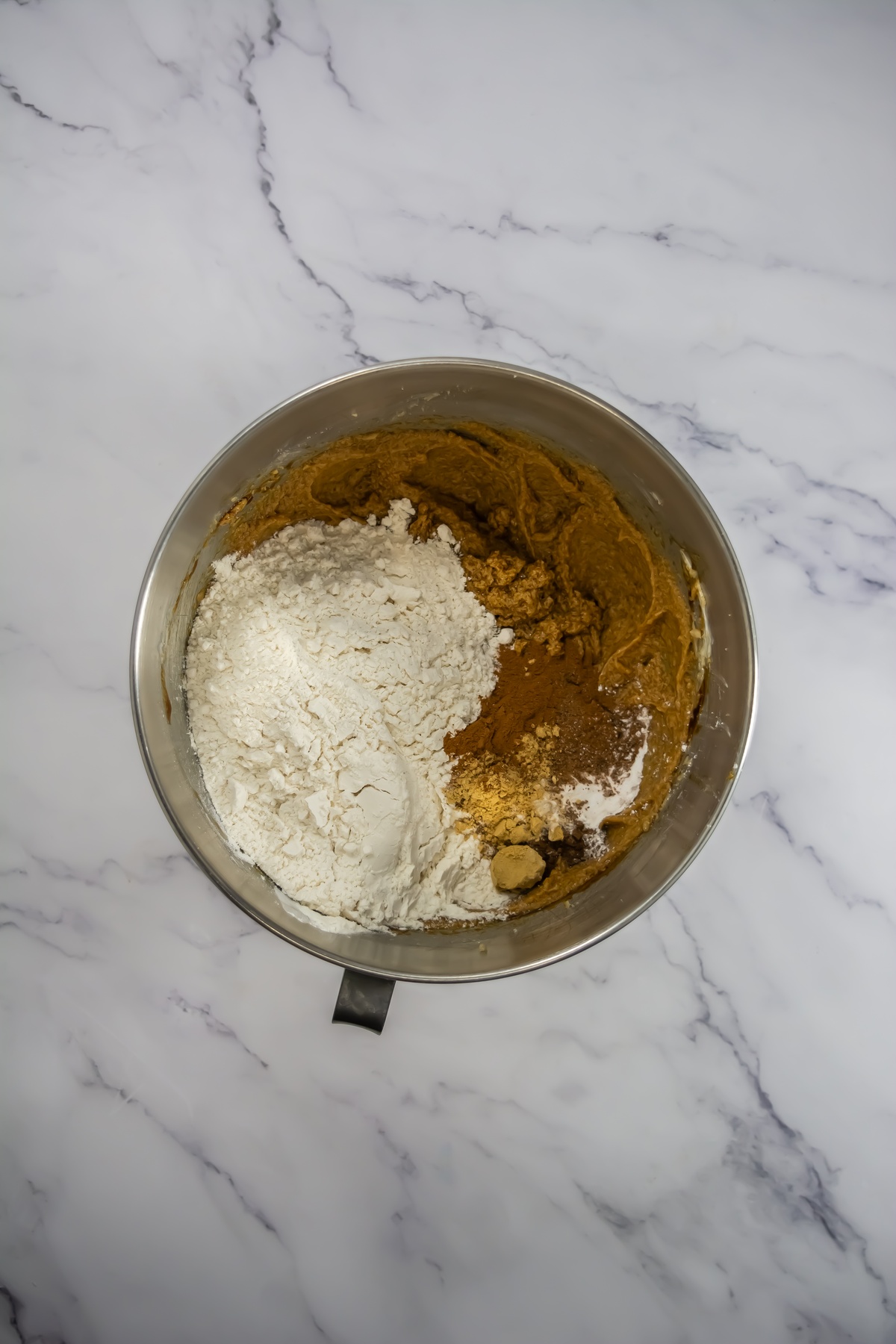 Stainless steel mixing bowl containing creamed brown batter with flour and spices added on top, unmixed, on marble surface.