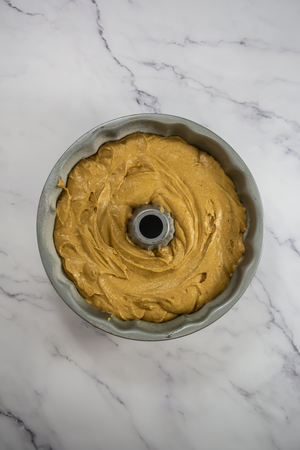 Golden-brown cake batter spread evenly in a greased bundt pan on a white marble surface, ready for baking.