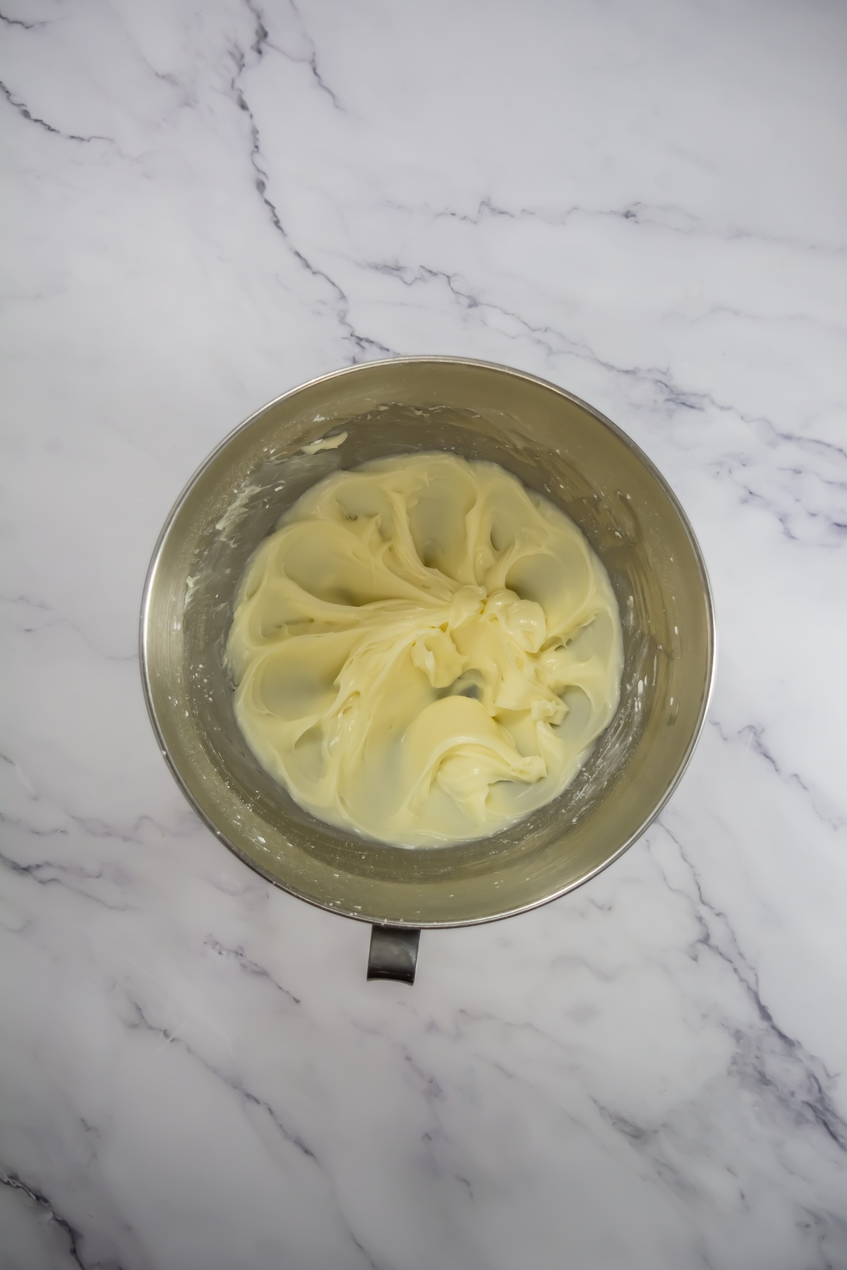 Creamy pale yellow frosting whipped to smooth peaks in a stainless steel mixing bowl on marble surface.