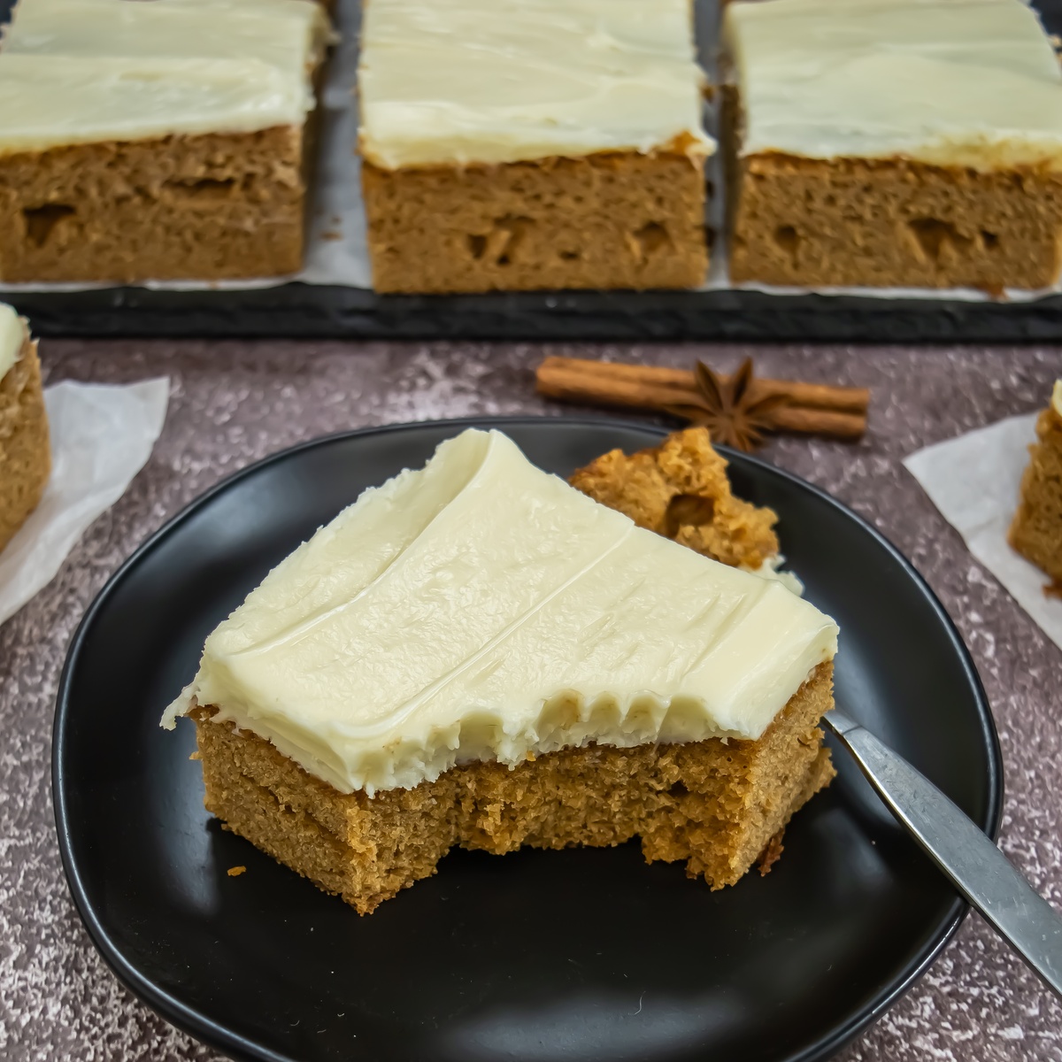 A slice of gingerbread sheet cake with cream cheese frosting on a black plate.