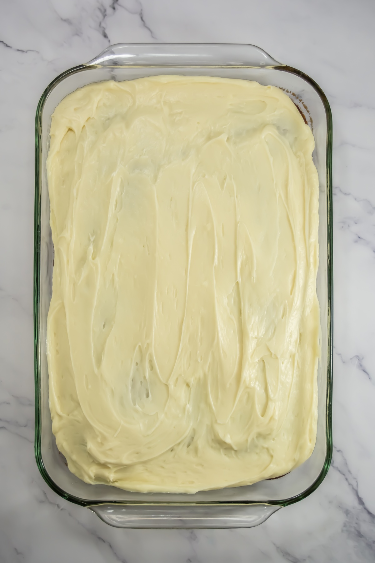 Gingerbread sheet cake topped with smooth cream cheese frosting in a glass 9x13 baking dish on a marble surface.