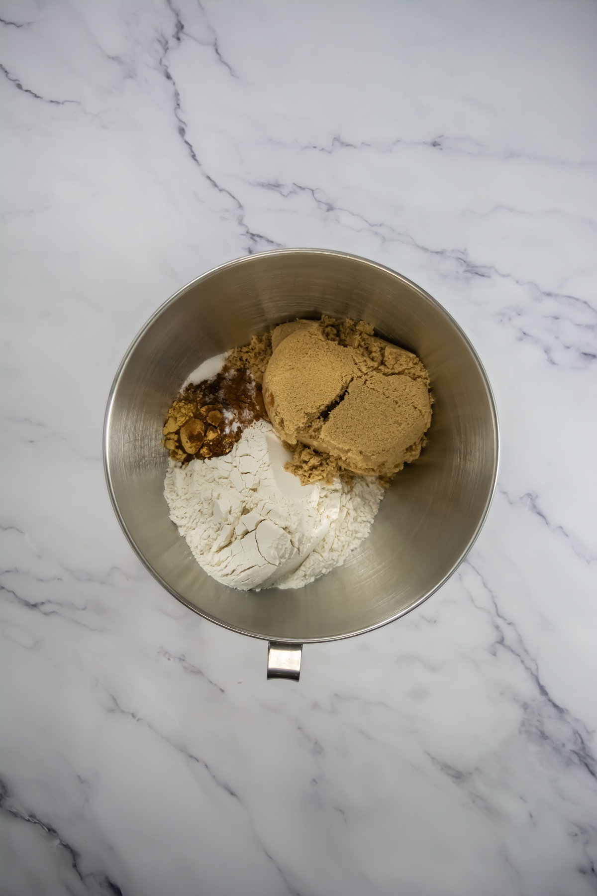 Stainless steel mixing bowl with unmixed flour, packed brown sugar, and spices on a marble surface.