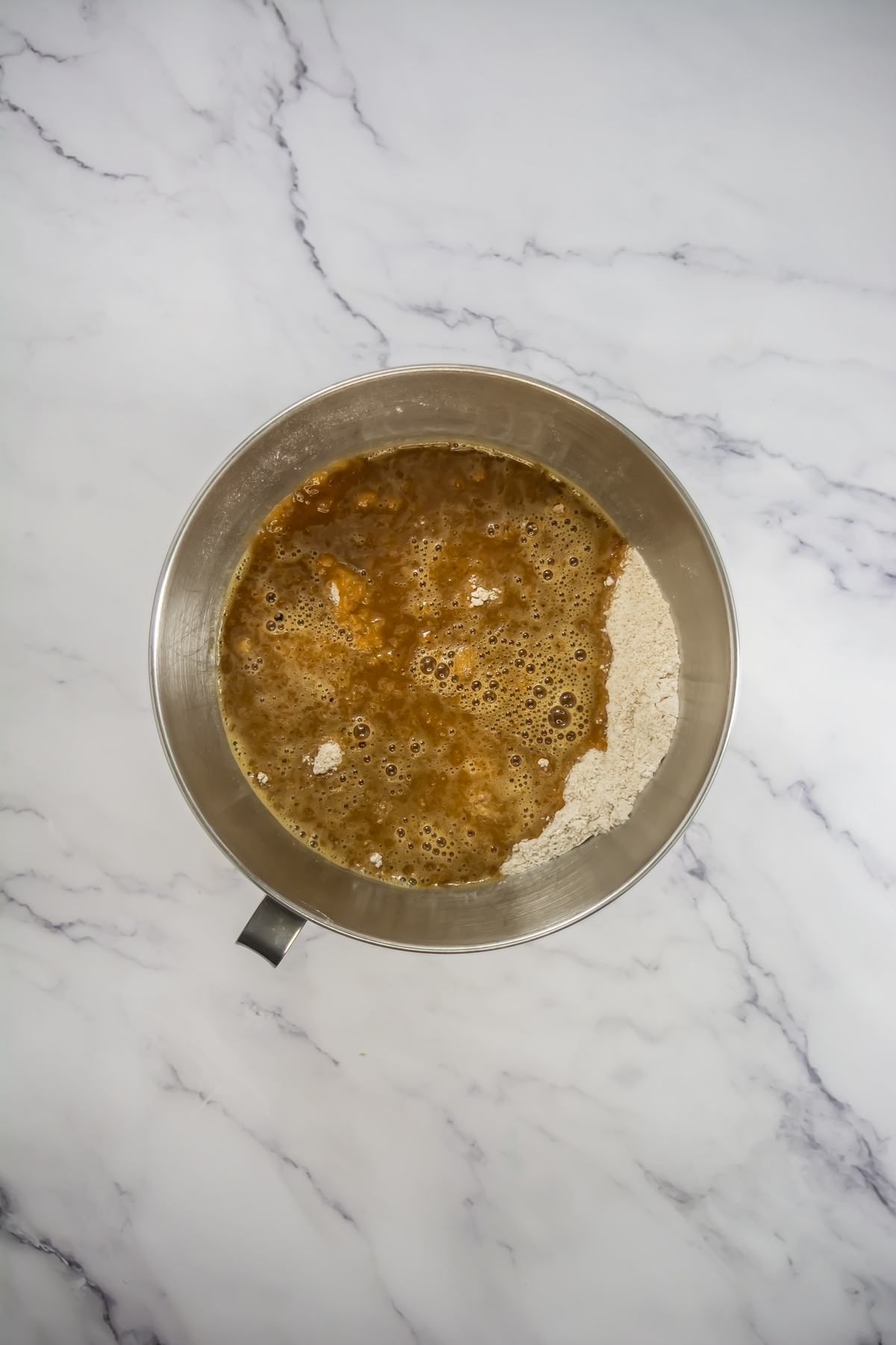 Wet ingredients with bubbles poured over dry flour in a stainless steel mixing bowl, not yet mixed together.