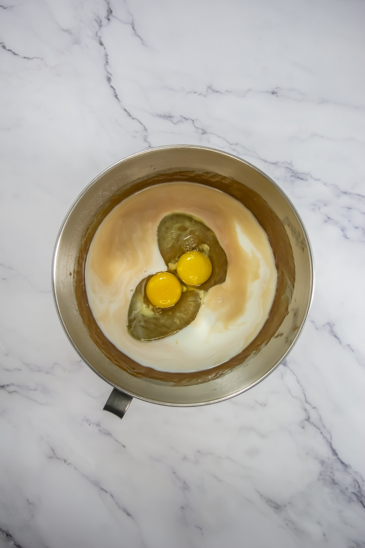 Two raw eggs cracked into a stainless steel mixing bowl with milk, ready to be mixed.
