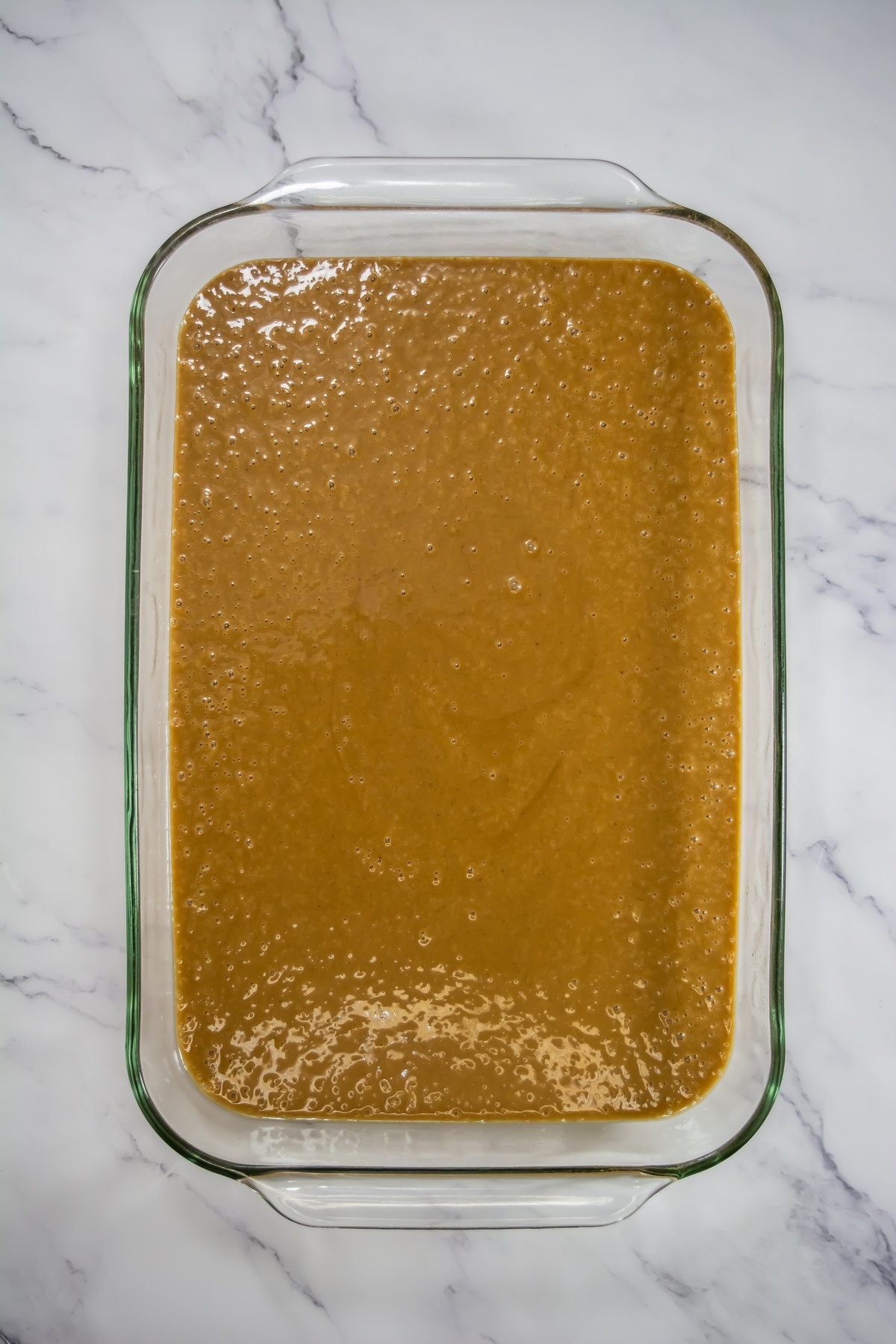Golden-brown gingerbread cake batter with small bubbles poured into a rectangular glass baking dish, ready to bake.