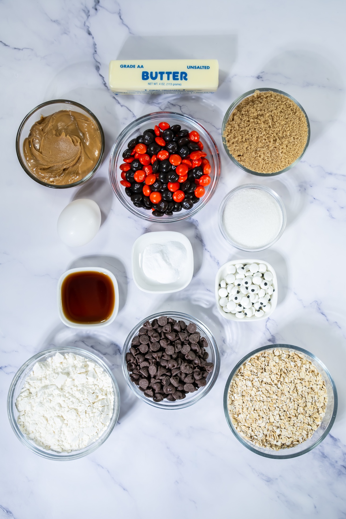 Ingredients for Halloween monster cookies: butter, peanut butter, egg, brown and white sugar, M&Ms, vanilla, baking soda on a table.