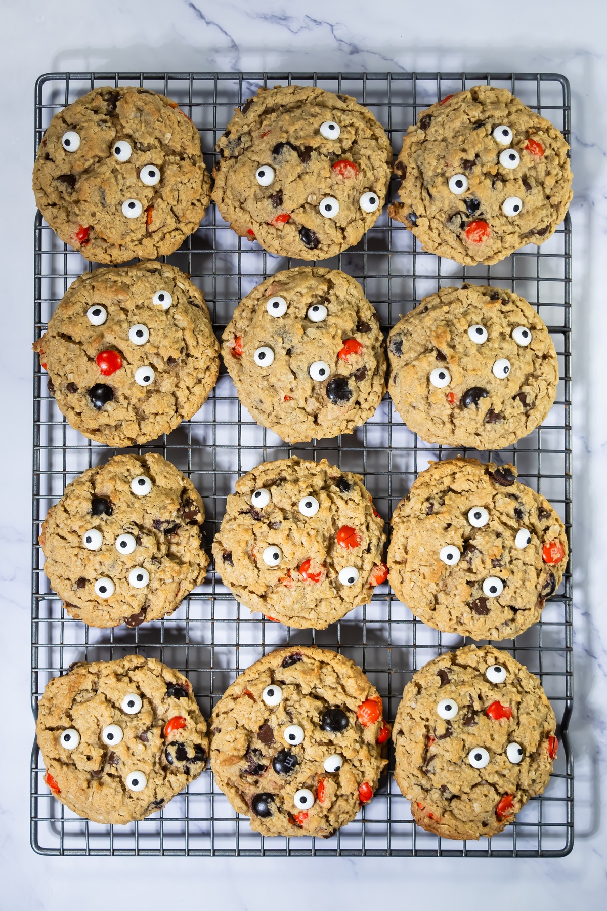 Twelve baked oatmeal cookies with candy eyes and orange and black candies cooling on a wire rack.