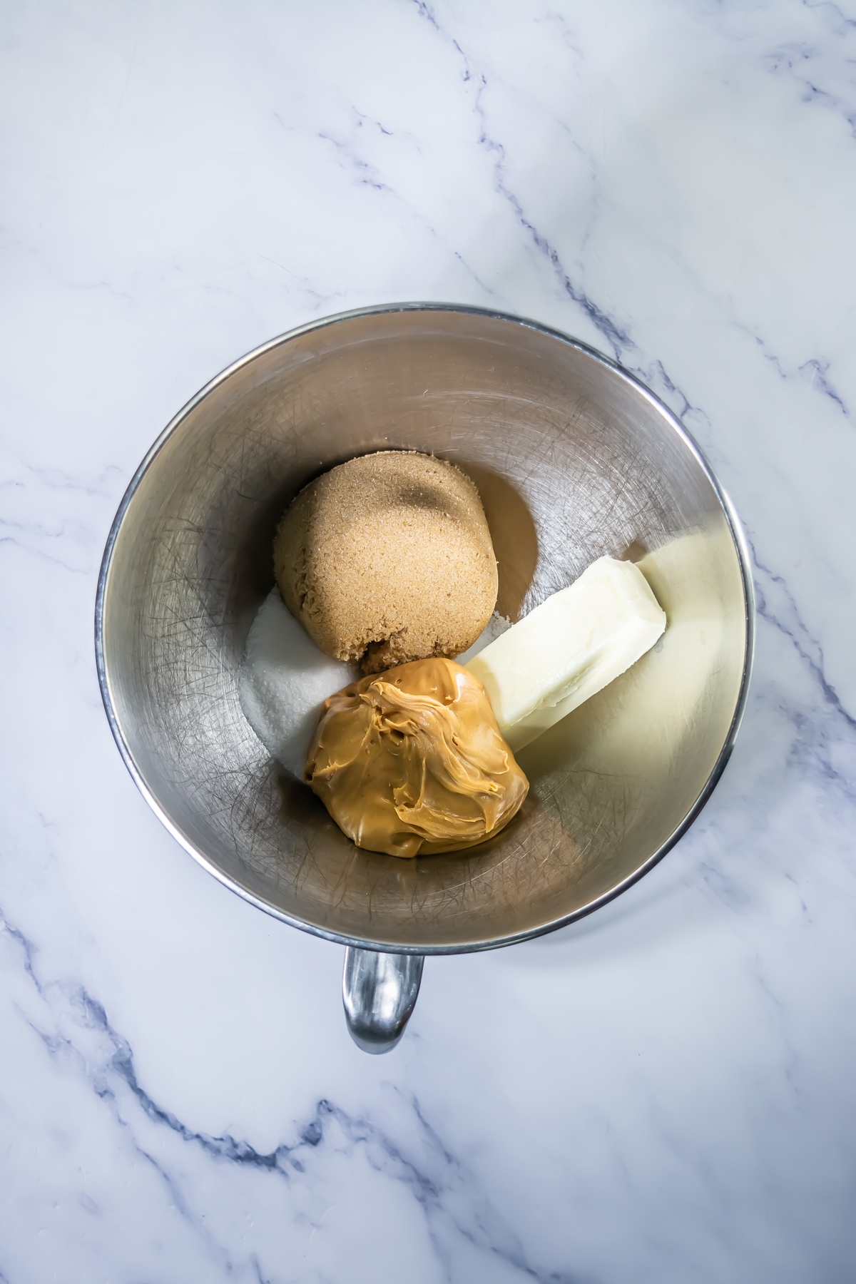 Stainless steel mixing bowl containing packed brown sugar, a stick of butter, and peanut butter before mixing.