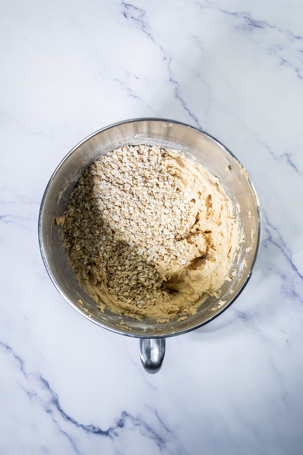 Rolled oats added to creamy cookie dough in a stainless steel mixing bowl on a marble surface, ready to be mixed.