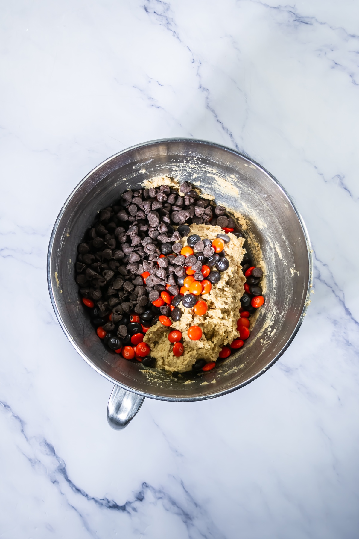 Cookie dough in a stainless steel mixing bowl with chocolate chips and orange and black M&M candies added, ready to mix.
