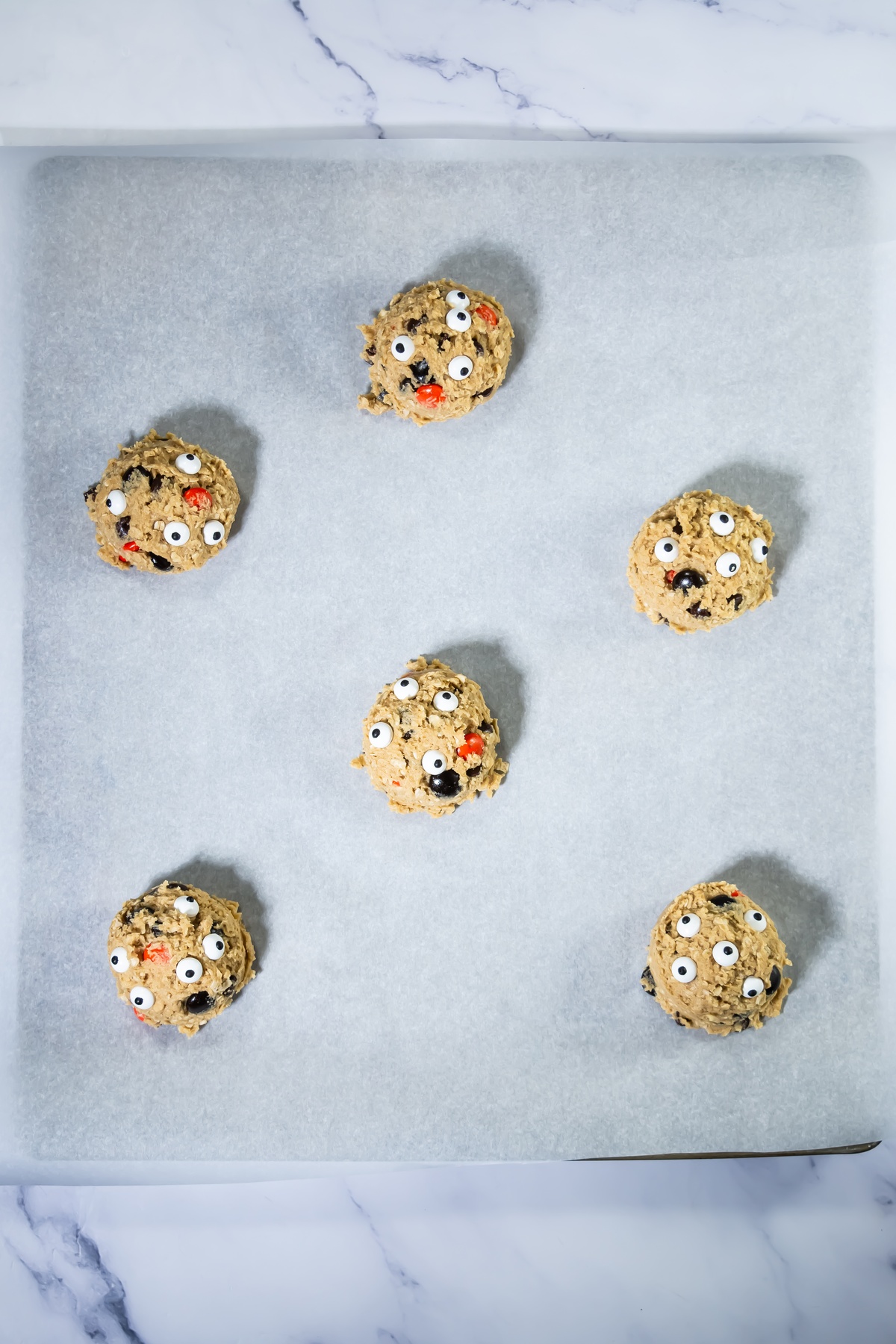 Seven unbaked cookie dough balls with candy eyes on a parchment-lined baking sheet, ready for the oven.