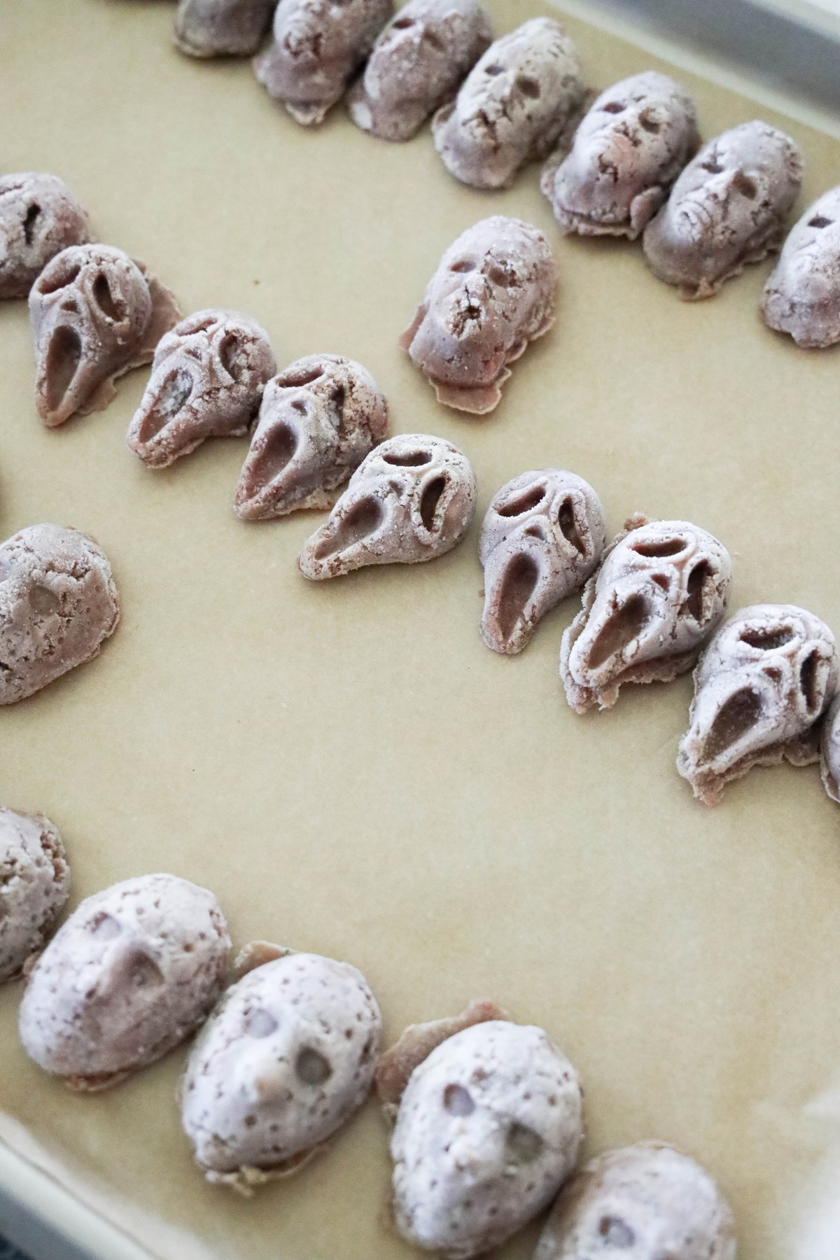 Rows of molded uncooked ghostface shaped meatballs arranged on parchment-lined baking sheet.