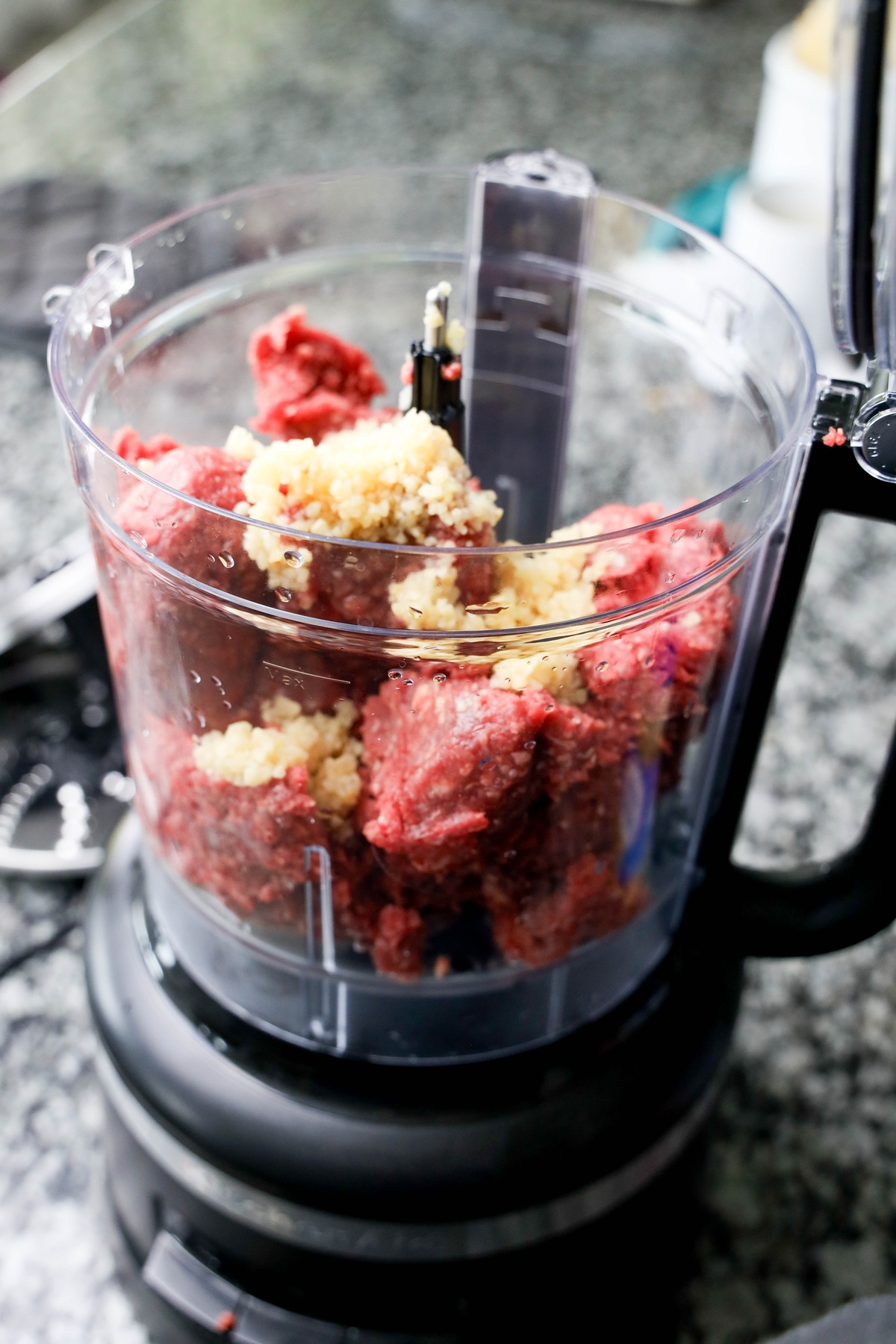 Raw ground beef and minced garlic in a food processor bowl, ready to be mixed.