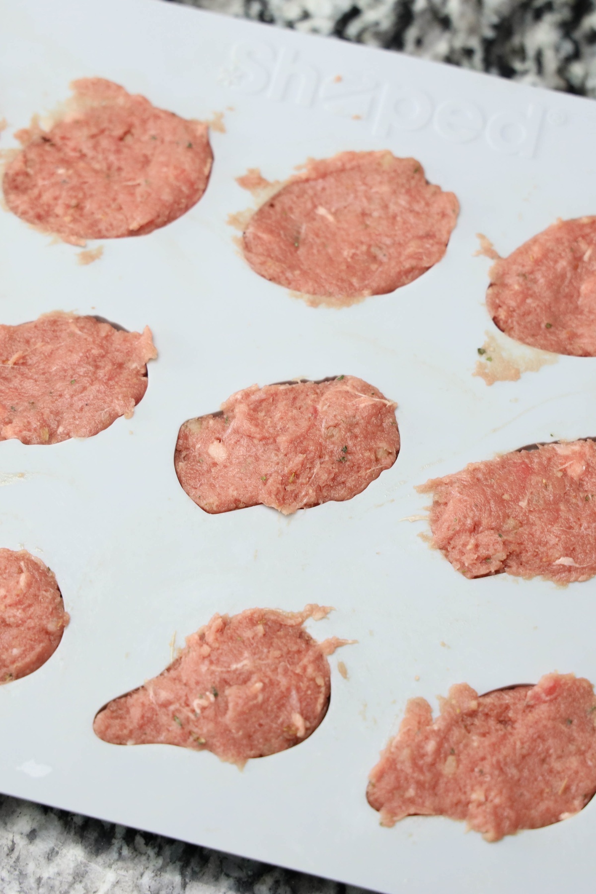 Raw seasoned ground meat patties shaped into irregular rounds on a white silicone mat on a granite countertop.