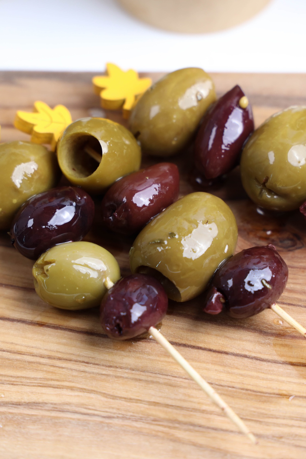Green and dark purple olives on wooden skewers with decorative yellow leaf picks, arranged on a wooden cutting board.