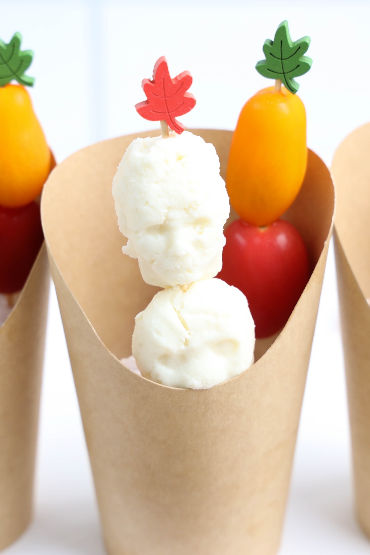 Skewers with mozzarella balls and cherry tomatoes topped with decorative leaf picks, displayed in brown paper cones.
