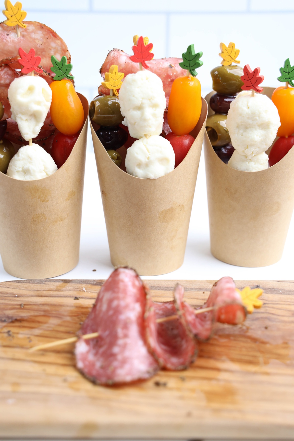 Charcuterie skewers with mozzarella balls, olives, cherry tomatoes, and salami in paper cups.