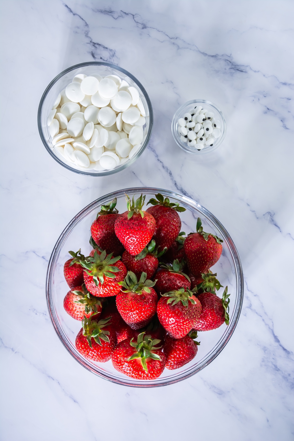 Fresh strawberries in a glass bowl, white candy melts, and candy eyes on a marble surface.