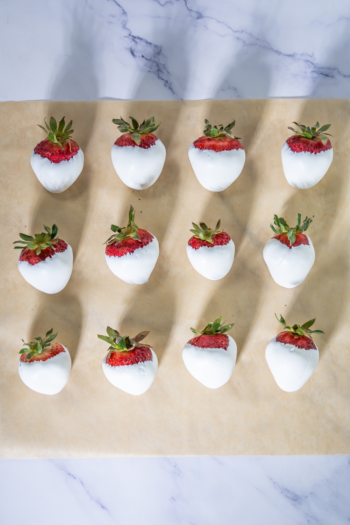 Twelve strawberries dipped in white chocolate arranged in rows on parchment paper, with green stems exposed at the top.