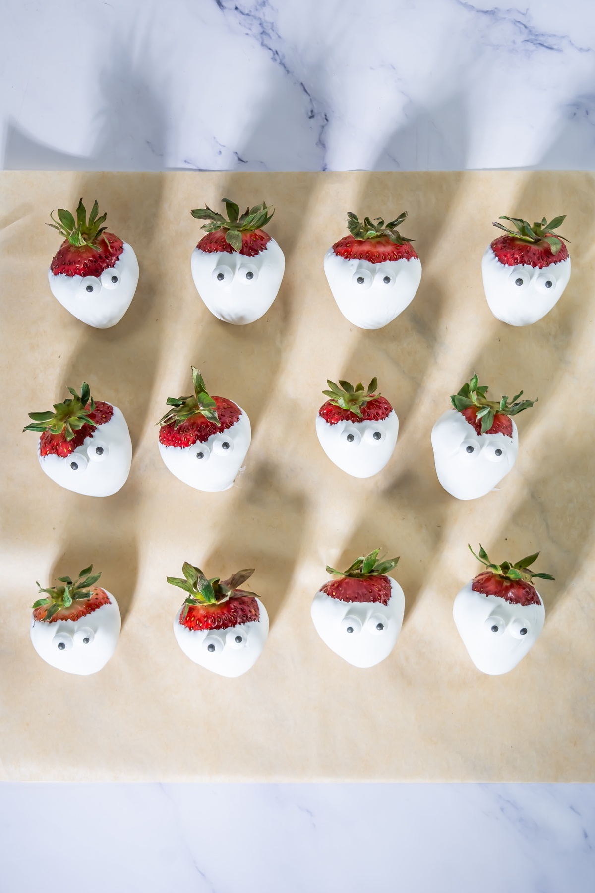 Twelve white chocolate-dipped strawberries with candy eyes arranged on parchment paper, styled as mummies for Halloween.
