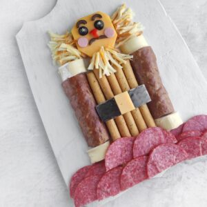 Charcuterie board arranged as a nutcracker with cheese face, shredded cheese hair, salami arms, breadstick body with belt.