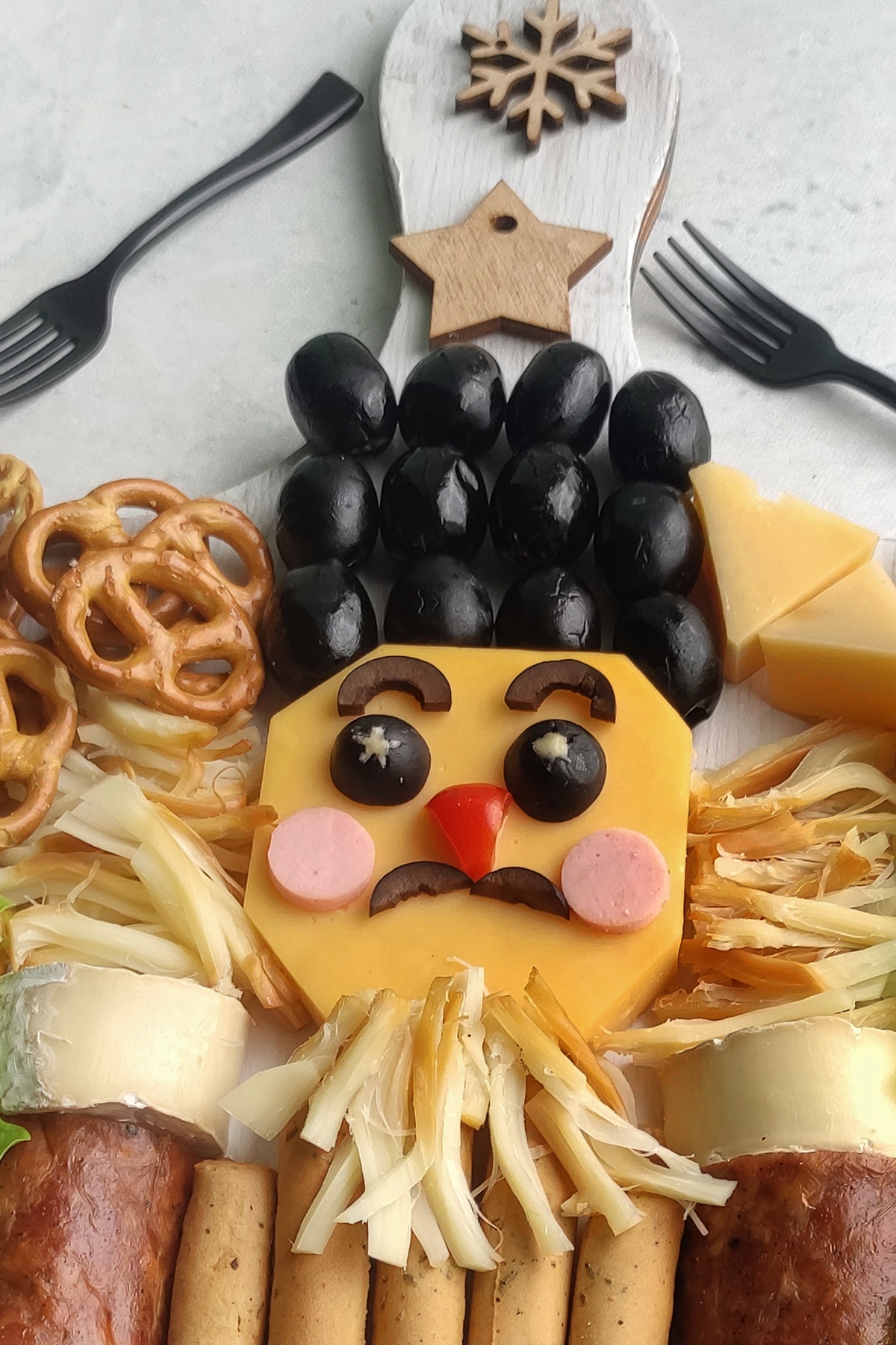 Nutcracker face made from cheese slice with black olive hair, olive eyes, red pepper nose, sausage cheeks.