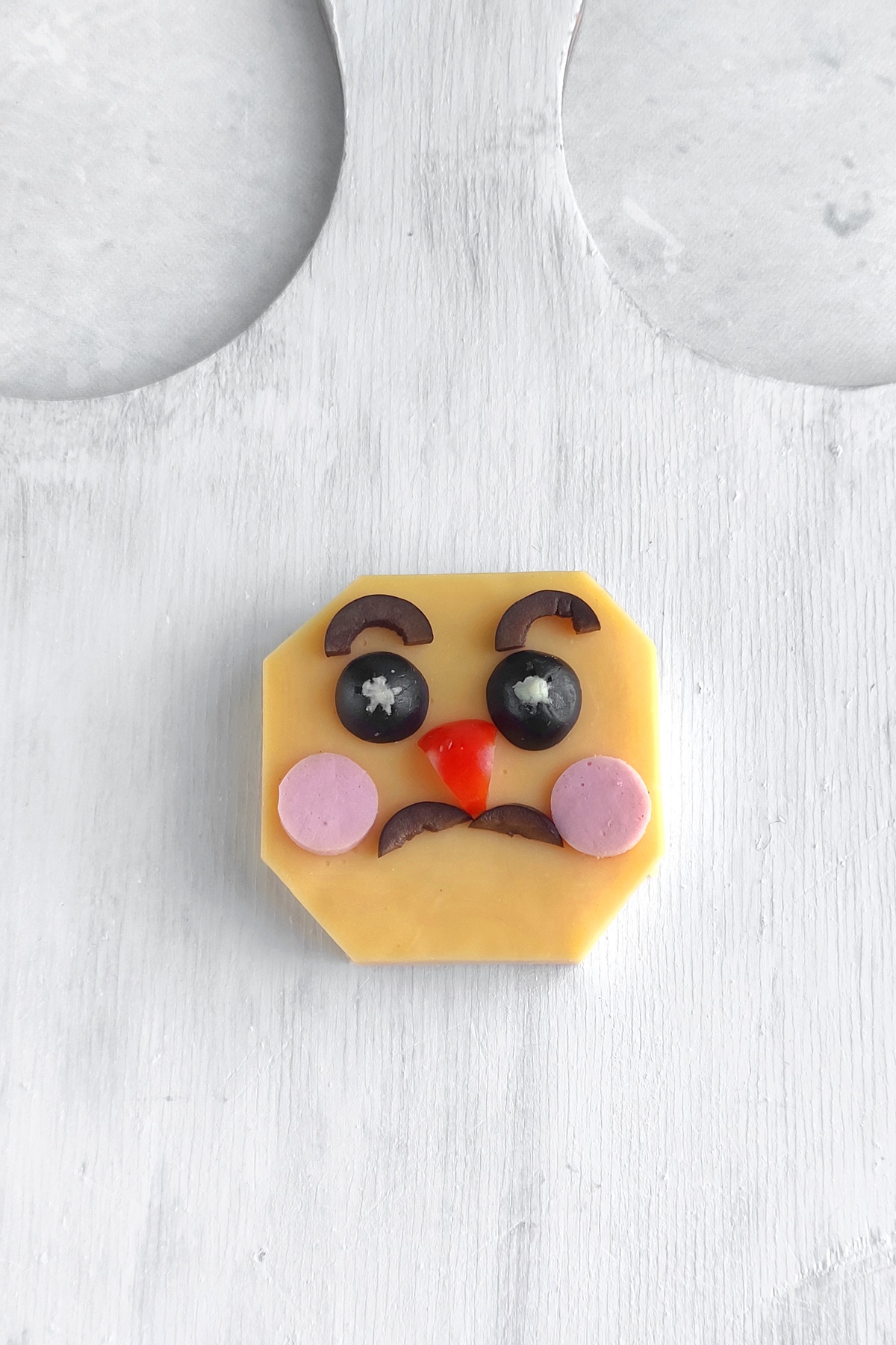 Cheese slice shaped into octagon with olive slices for eyes and eyebrows, red pepper nose, and pink circular cheeks.